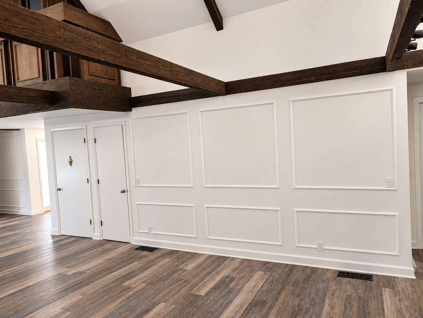 Empty room with white paneled walls, wooden flooring, and exposed dark wooden beams on the ceiling. There are two doors on the left, one with a decorative door knocker. Vent covers are on the floor.
