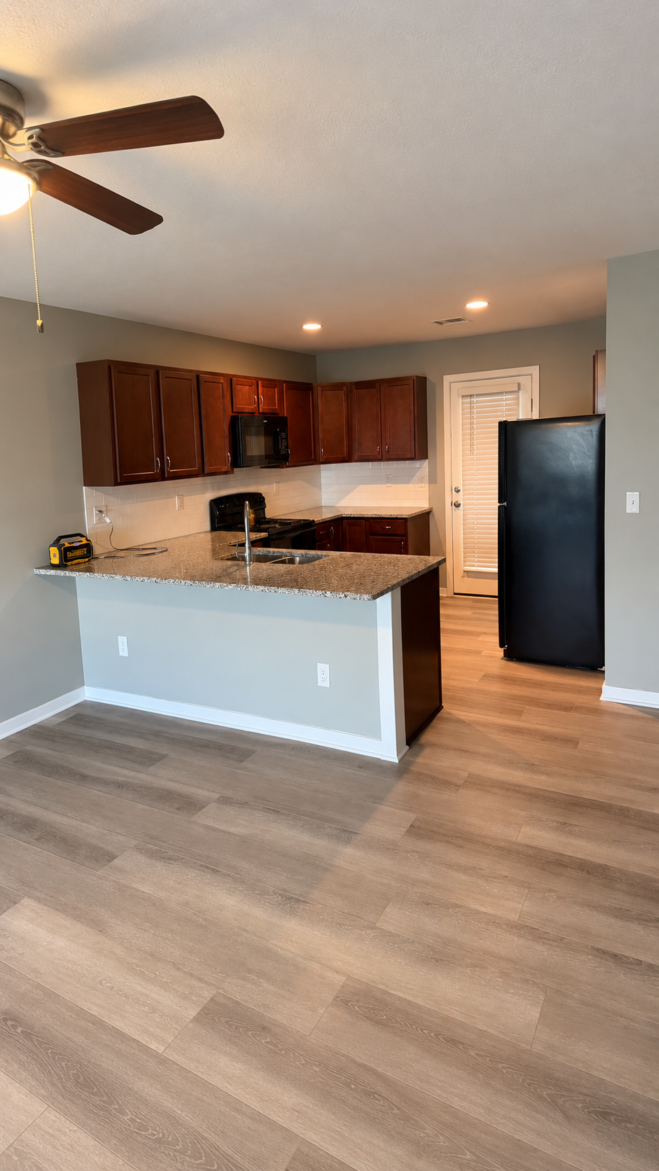 Empty kitchen with wooden cabinets, granite countertop, black refrigerator, black microwave, and a door with blinds, hardwood flooring, and ceiling fan.