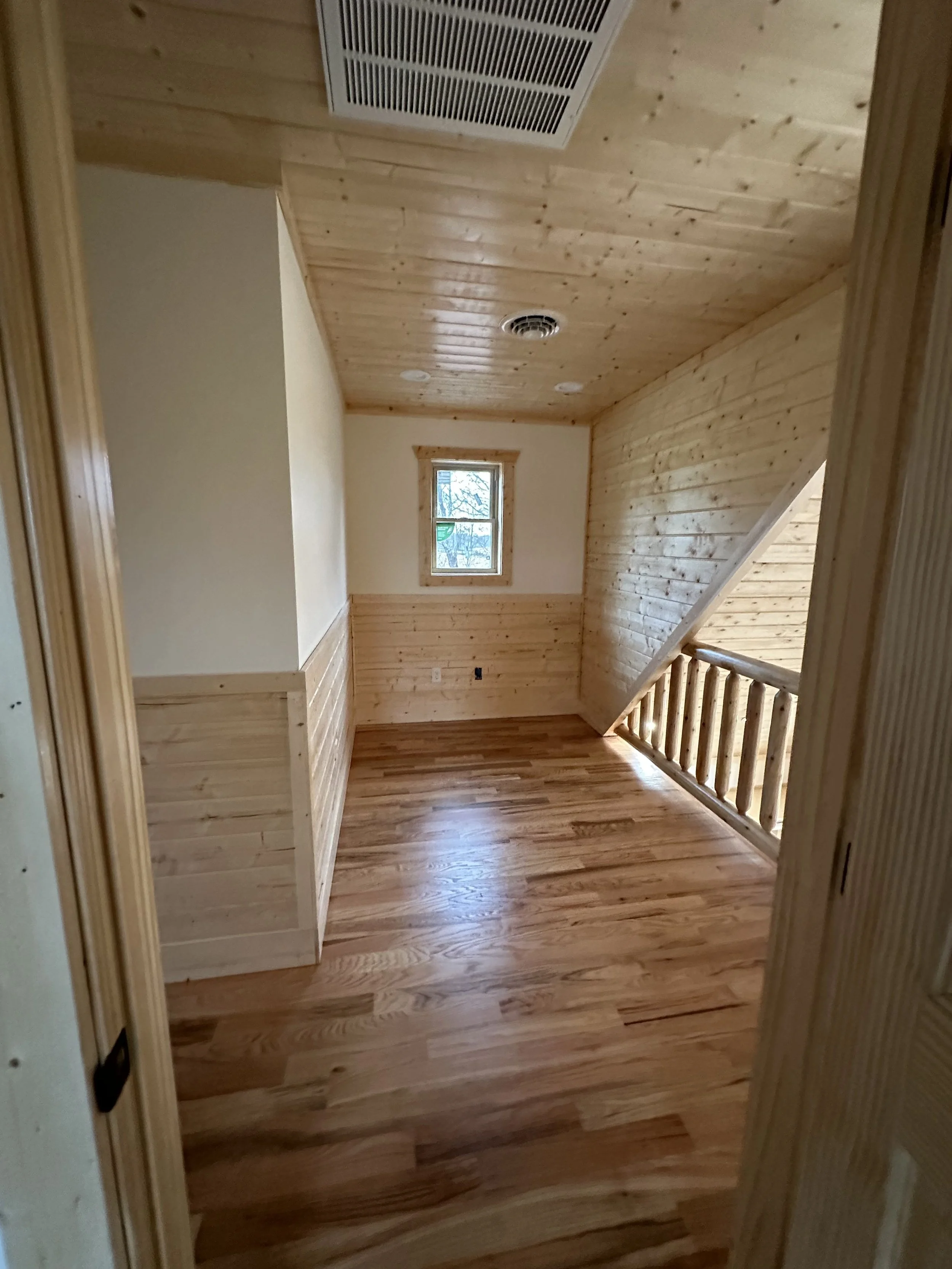 A small room with wood-paneled walls, a wood floor, a small window, and a staircase with a wooden railing. The ceiling has a vent and a circular smoke detector.
