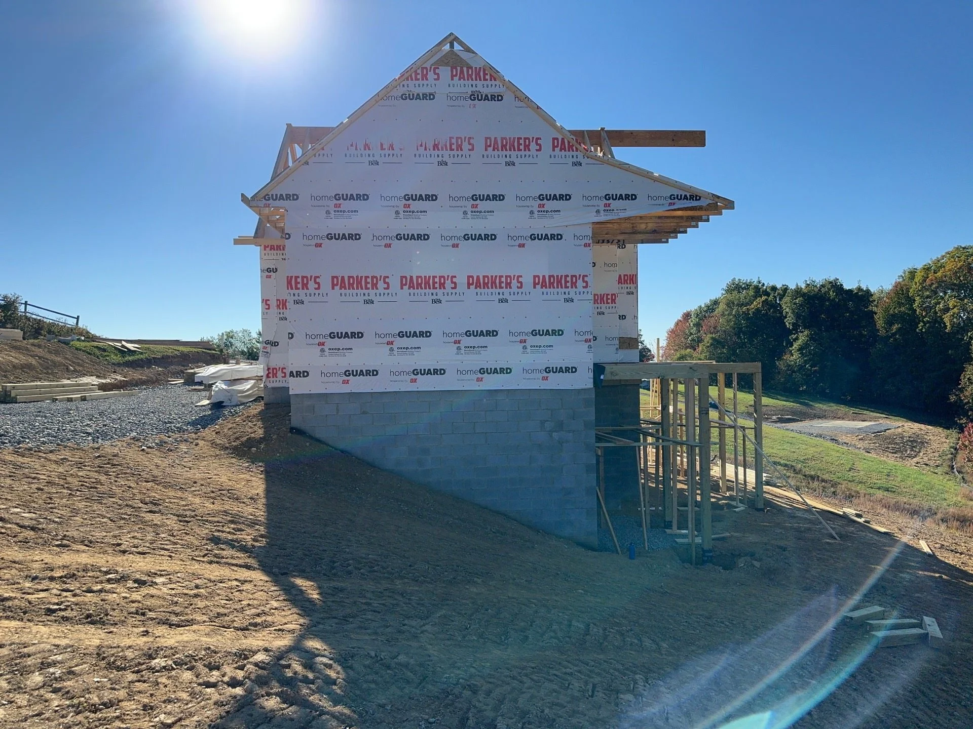 Side view of a house under construction with a partly built brick foundation and wooden framing, wrapped in building material, on a construction site with dirt and gravel ground, trees in the background, and a bright sunny sky.