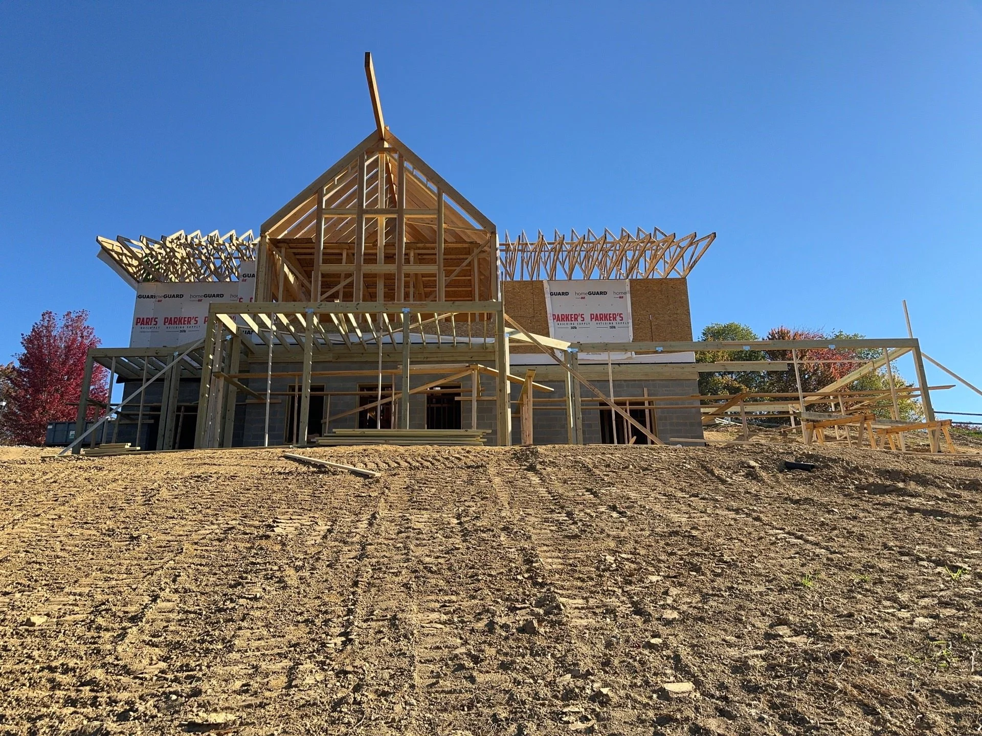 A house under construction with a wooden framework and scaffolding in front of a clear blue sky, with some trees visible in the background.
