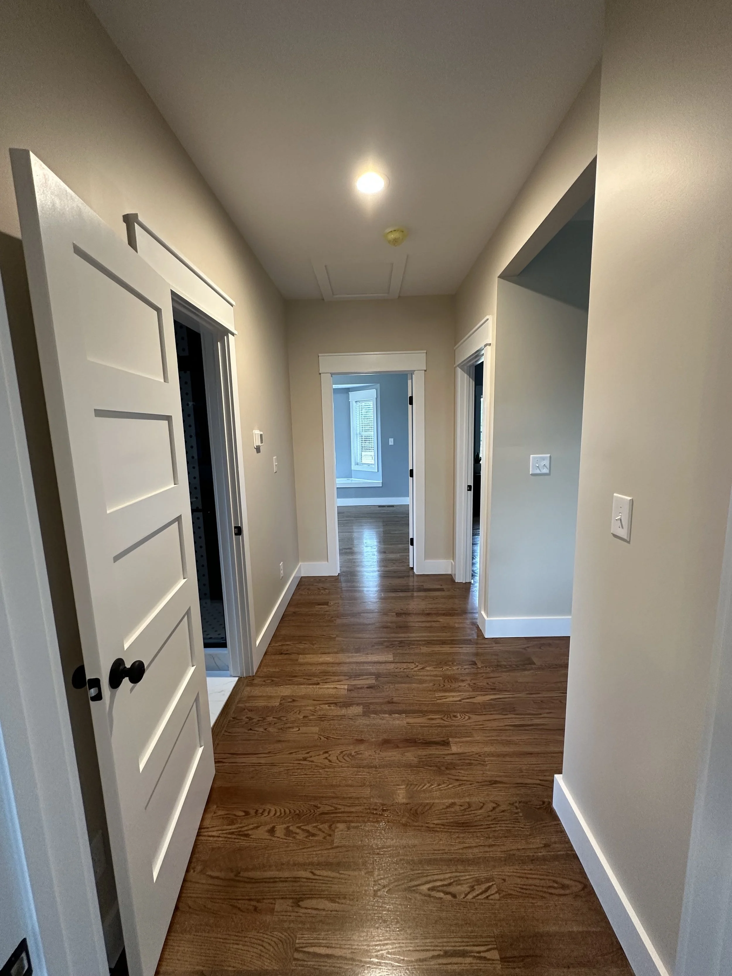 A hallway with hardwood floors, beige walls, white door frames, and a ceiling light. Two open doors on the left and right, leading to other rooms, and an open doorway at the end showing a room with a window.