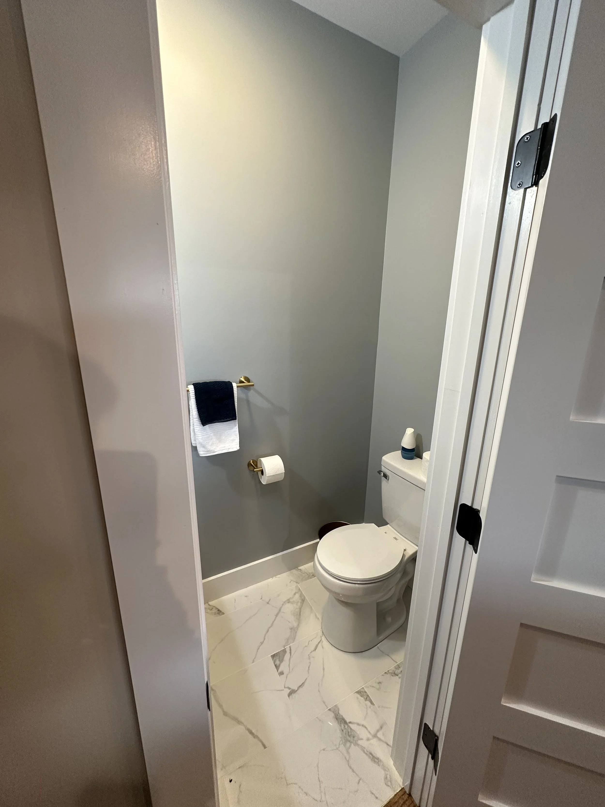 A small bathroom with a white toilet, a gold towel bar with a navy and white towel, a toilet paper roll, and a marble floor. The wall behind the toilet is painted a light gray color.