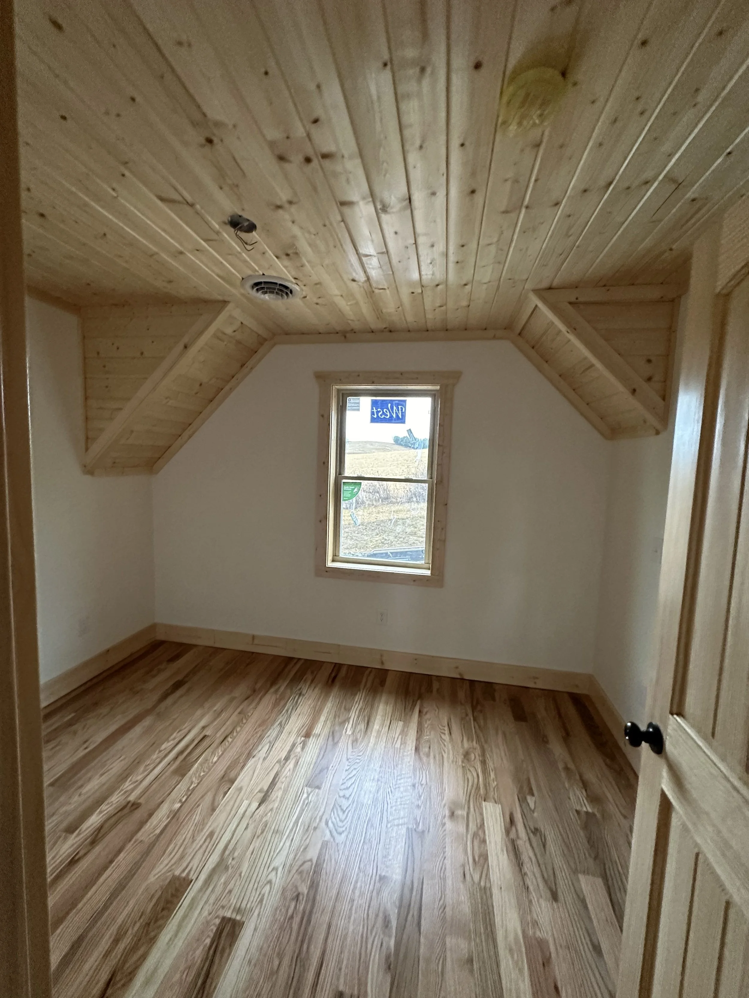A small bedroom with wooden walls, ceiling, and floor. There is a single window on the back wall, and the room appears to be lit by natural light coming through the window.