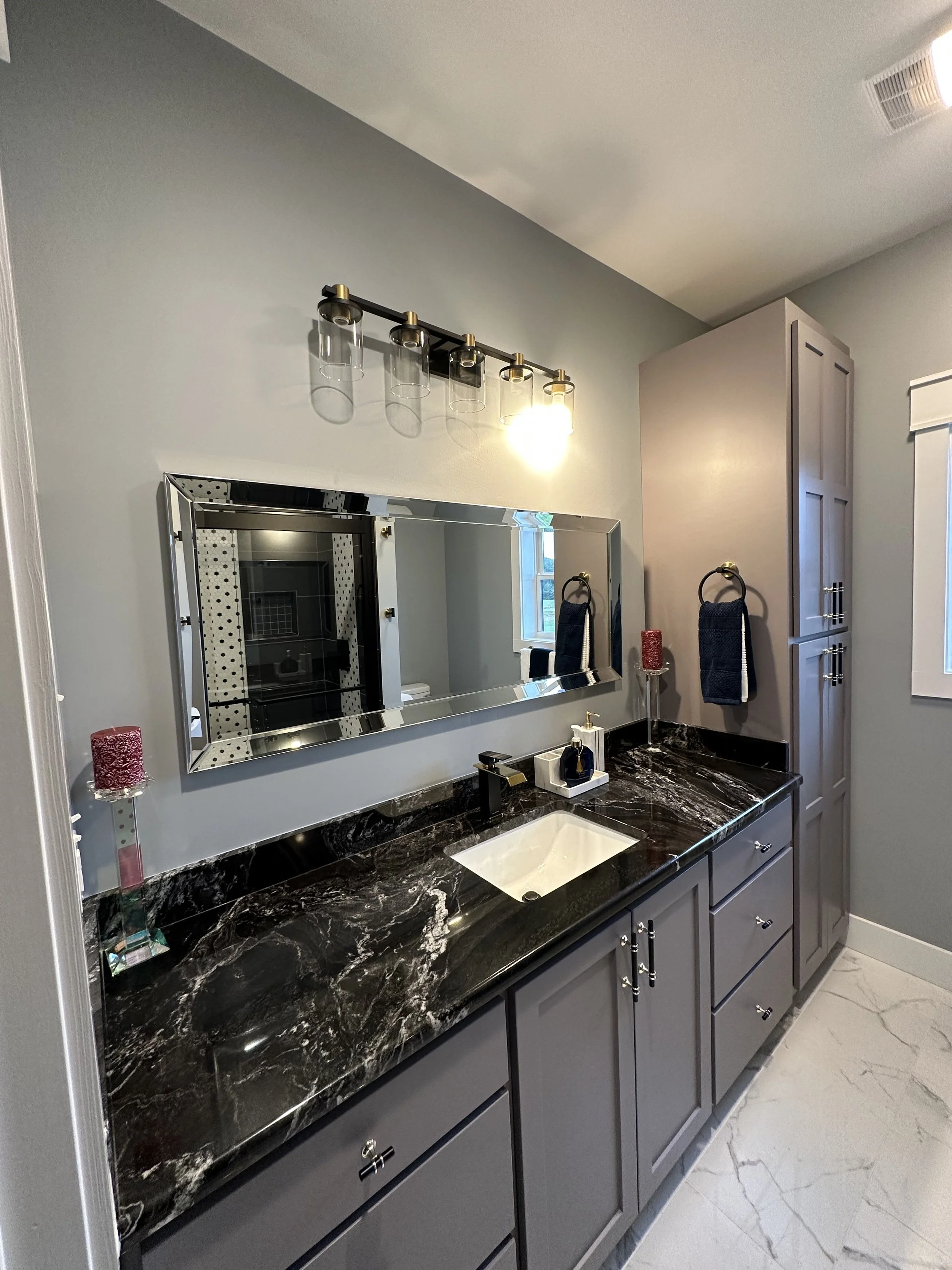 A modern bathroom vanity with a black marble countertop, gray cabinets, and a large mirror. There are two navy blue towels hanging on the wall and some decorative items on the countertop. A light fixture with five glass shades is mounted above the mi