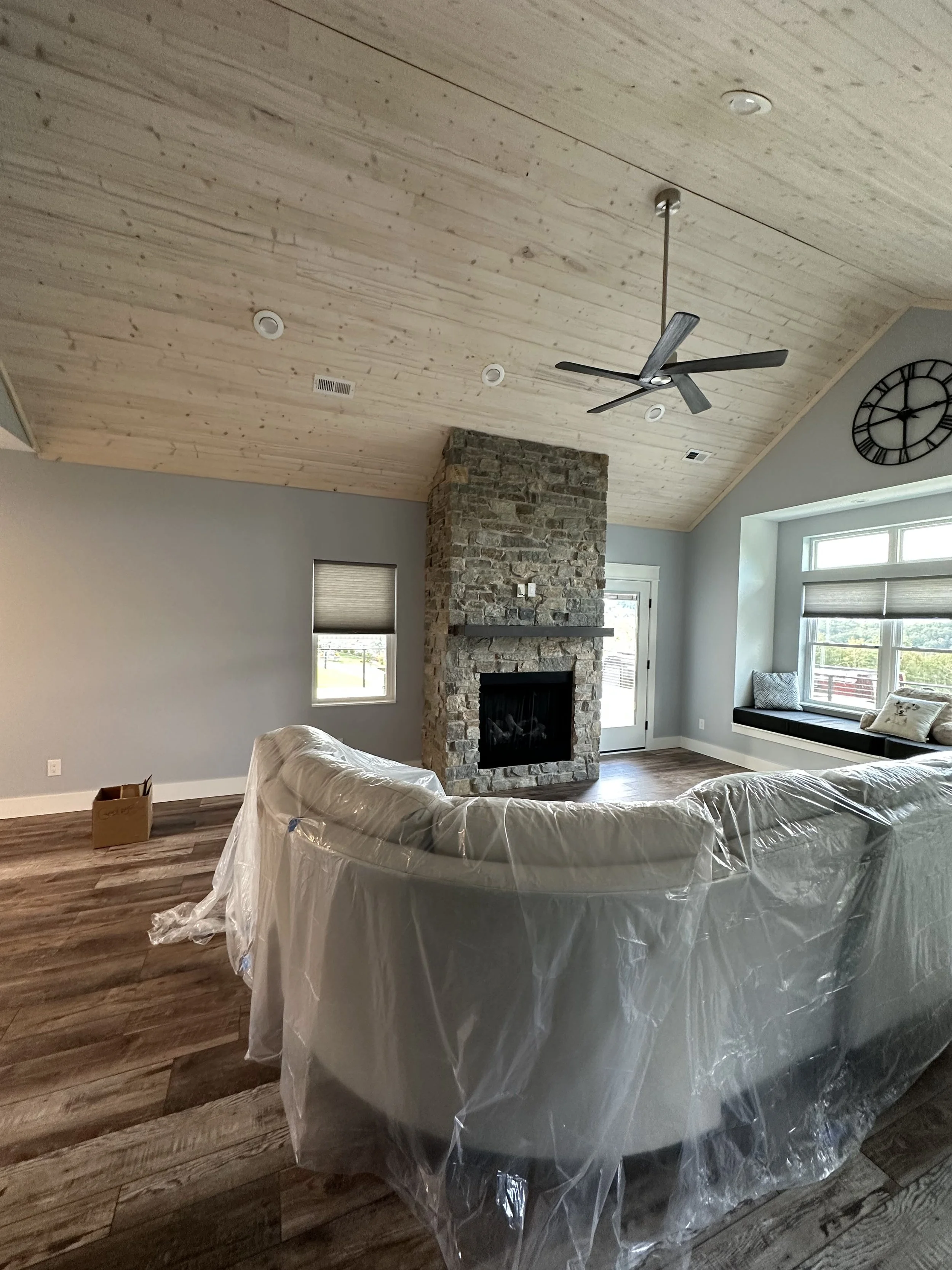 Living room with a wood-paneled ceiling, stone fireplace, window bench with pillows, and plastic-covered furniture.