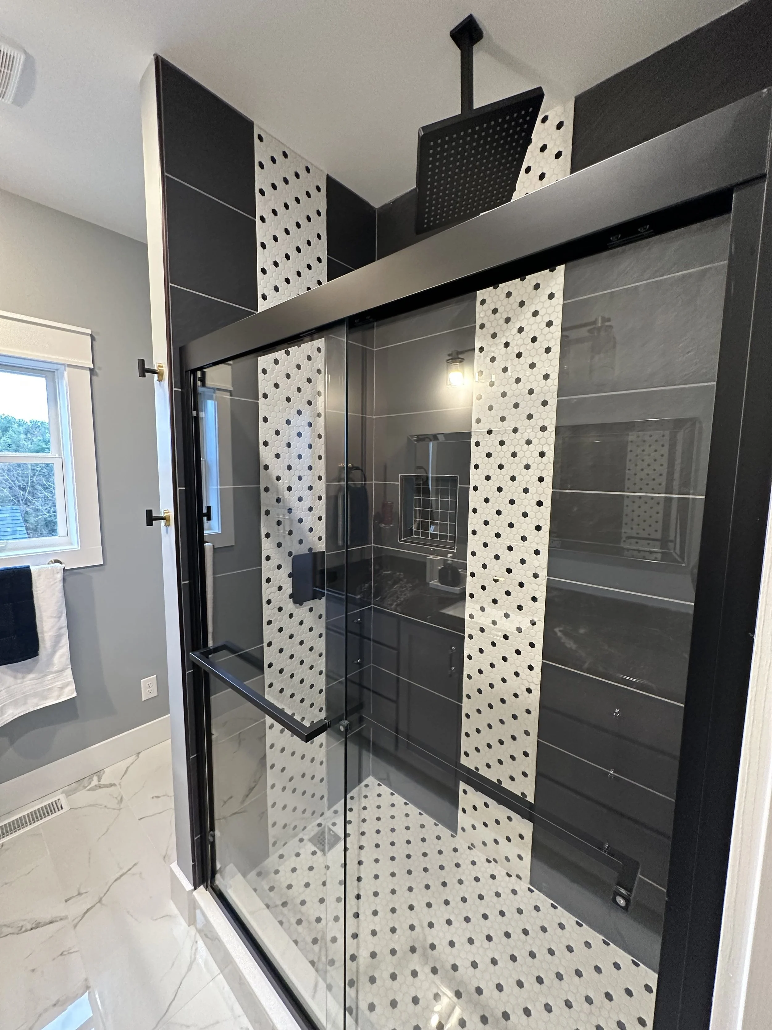 Modern walk-in shower with black and white mosaic tile accents, glass door, black shower head, gray walls, and white marble floor outside the shower.