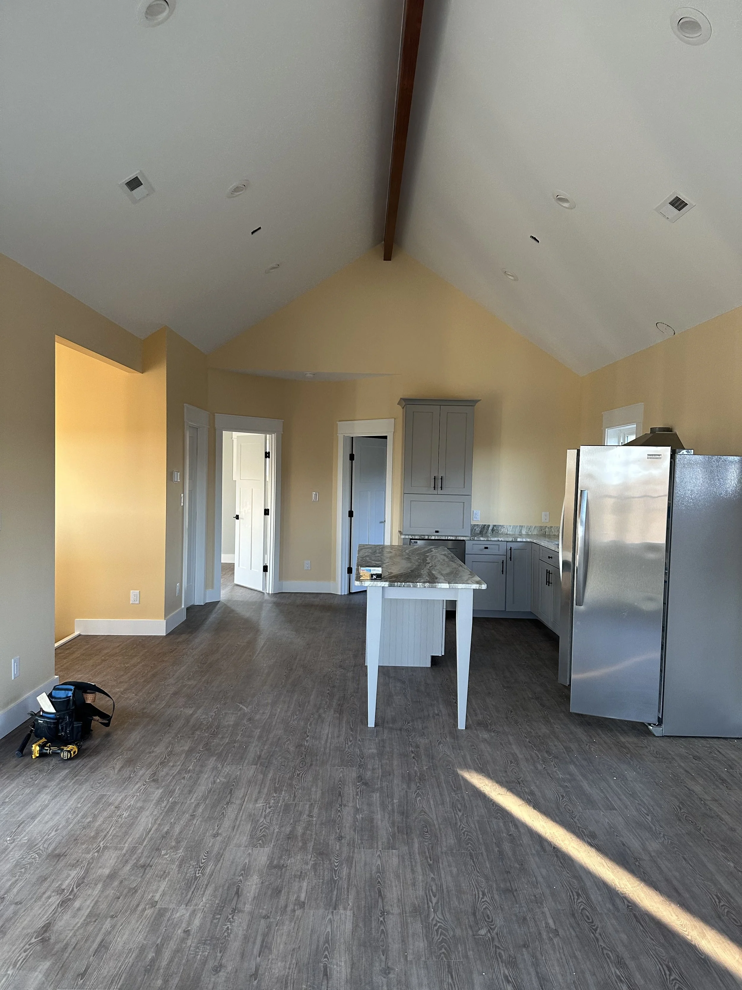 Interior view of a kitchen with light yellow walls, gray cabinets, and a marble countertop island. Stainless steel refrigerator on the right, and three doorways visible in the background. The ceiling has several fixtures and a wooden beam.