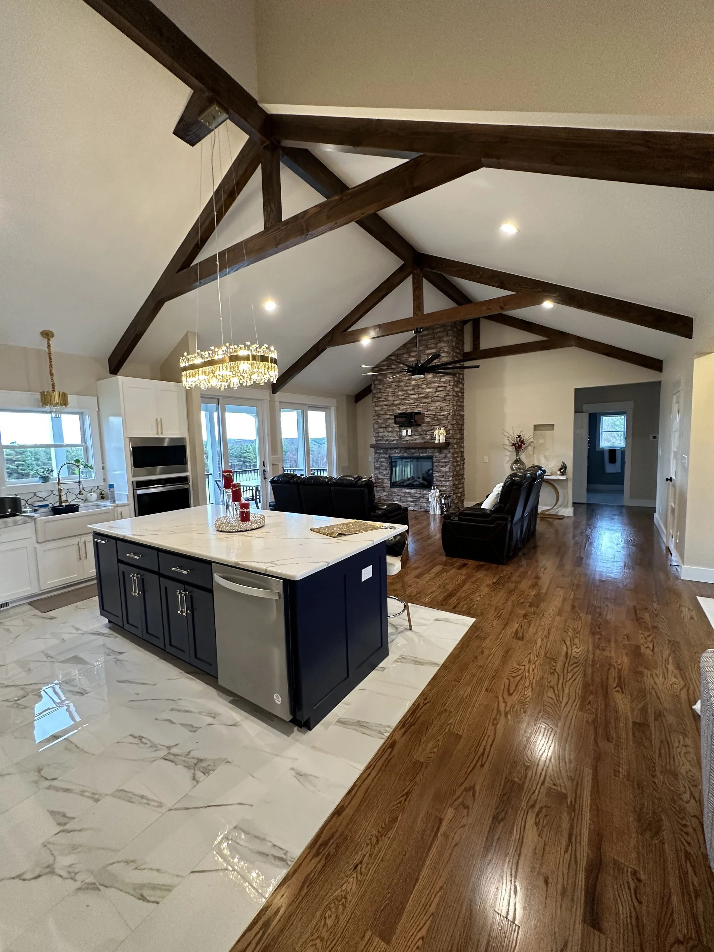 A spacious kitchen and living room area with vaulted ceilings, exposed wooden beams, a fireplace with a stone surround, and a large island with a marble top. The room is well-lit with windows, a chandelier, and recessed ceiling lights. The flooring f