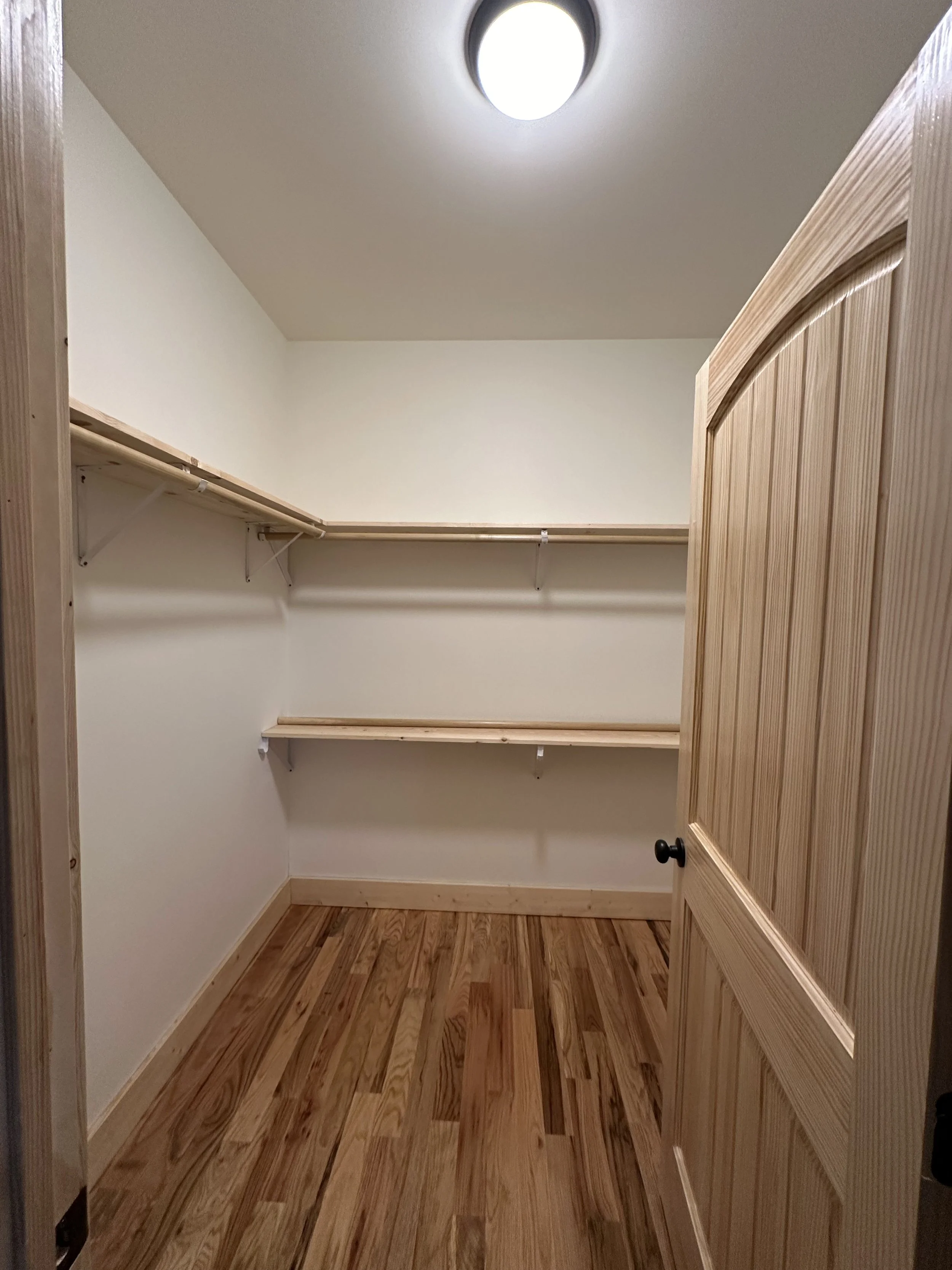 Empty walk-in closet with wooden shelves and a hardwood floor, with a ceiling light fixture.