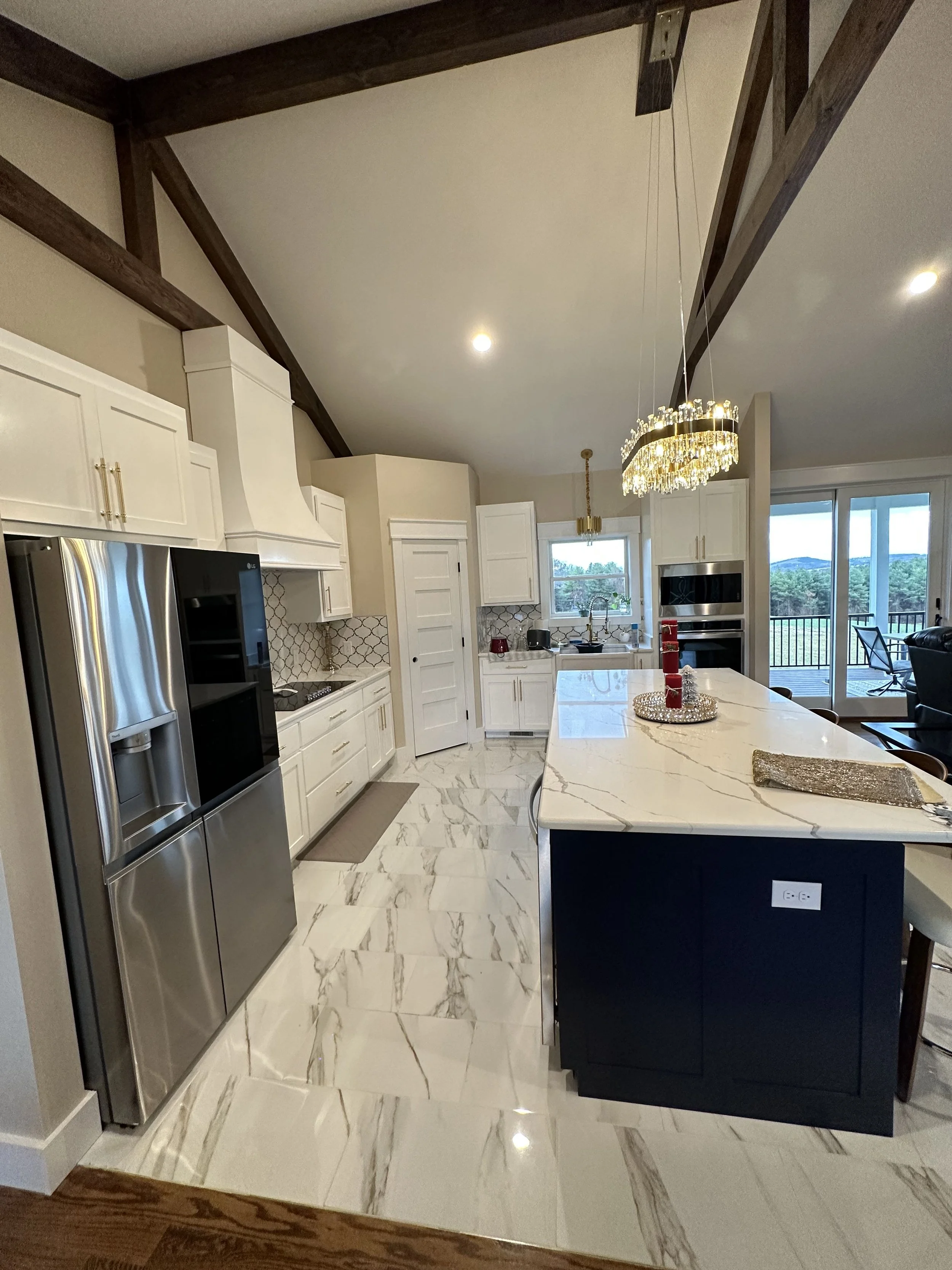 Modern kitchen with white cabinets, stainless steel refrigerator, marble island, and glass sliding door leading to a porch. Hanging chandelier and natural light from windows.