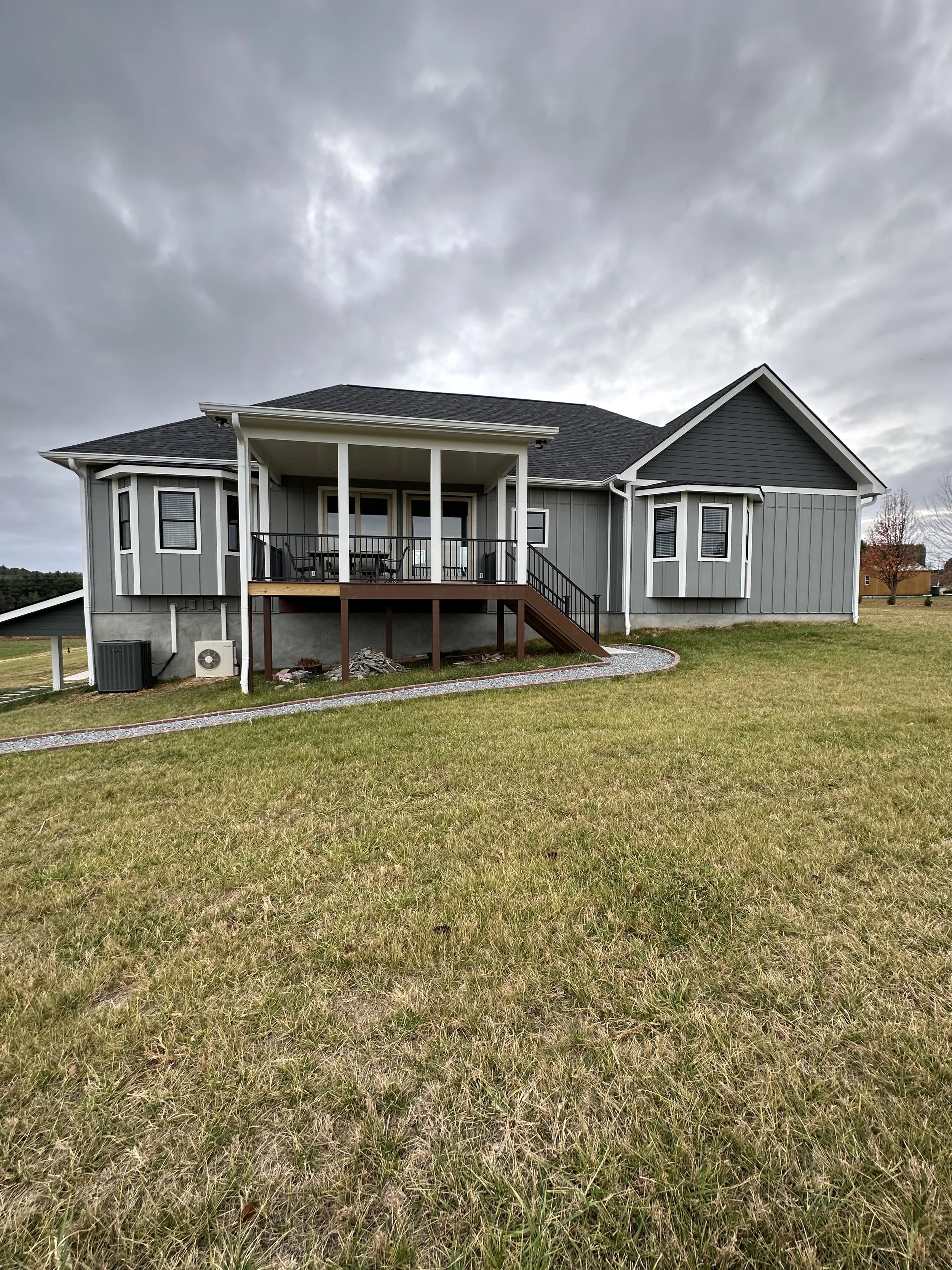 The image depicts the back of a gray house with a small porch, stairs, and railing. The house has several windows and a roof with dark shingles. The lawn in front of the house is green, and there are trees and other houses in the background. The sky above is cloudy and overcast.