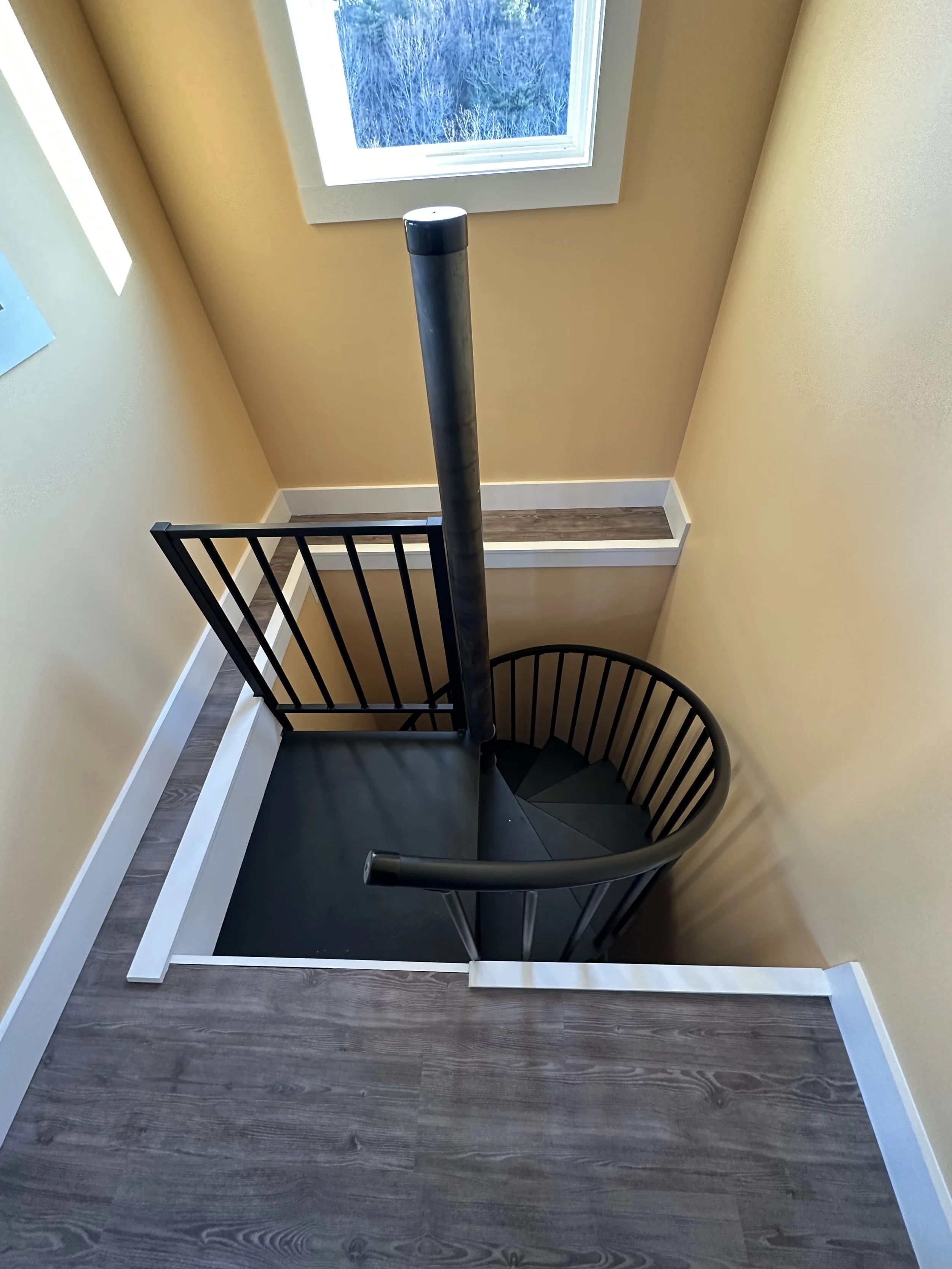 Top view of a spiral staircase with black handrails, surrounded by beige walls and a window showing an outdoor scene of trees and sky.
