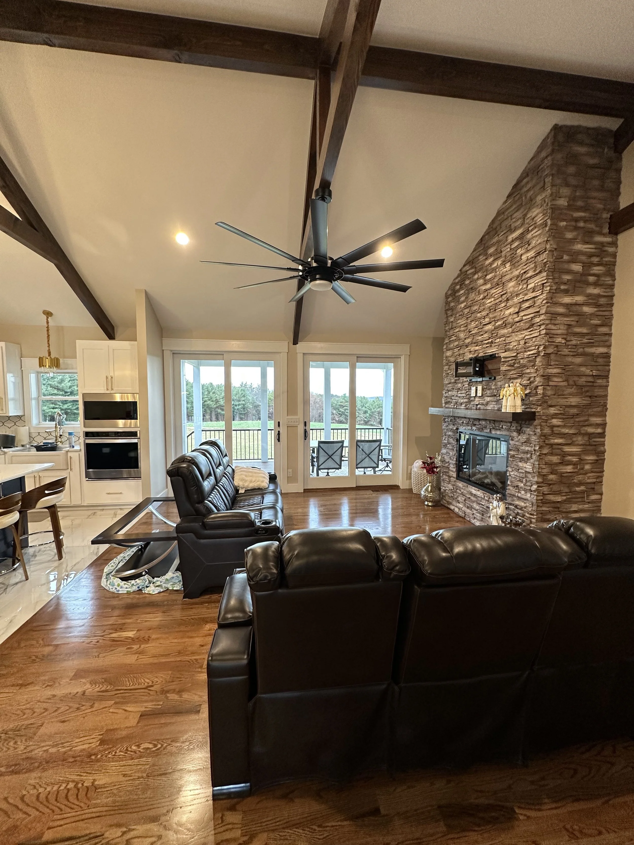 Living room with a high ceiling, ceiling fan, stone fireplace, black leather furniture, and a view of a deck and trees outside.