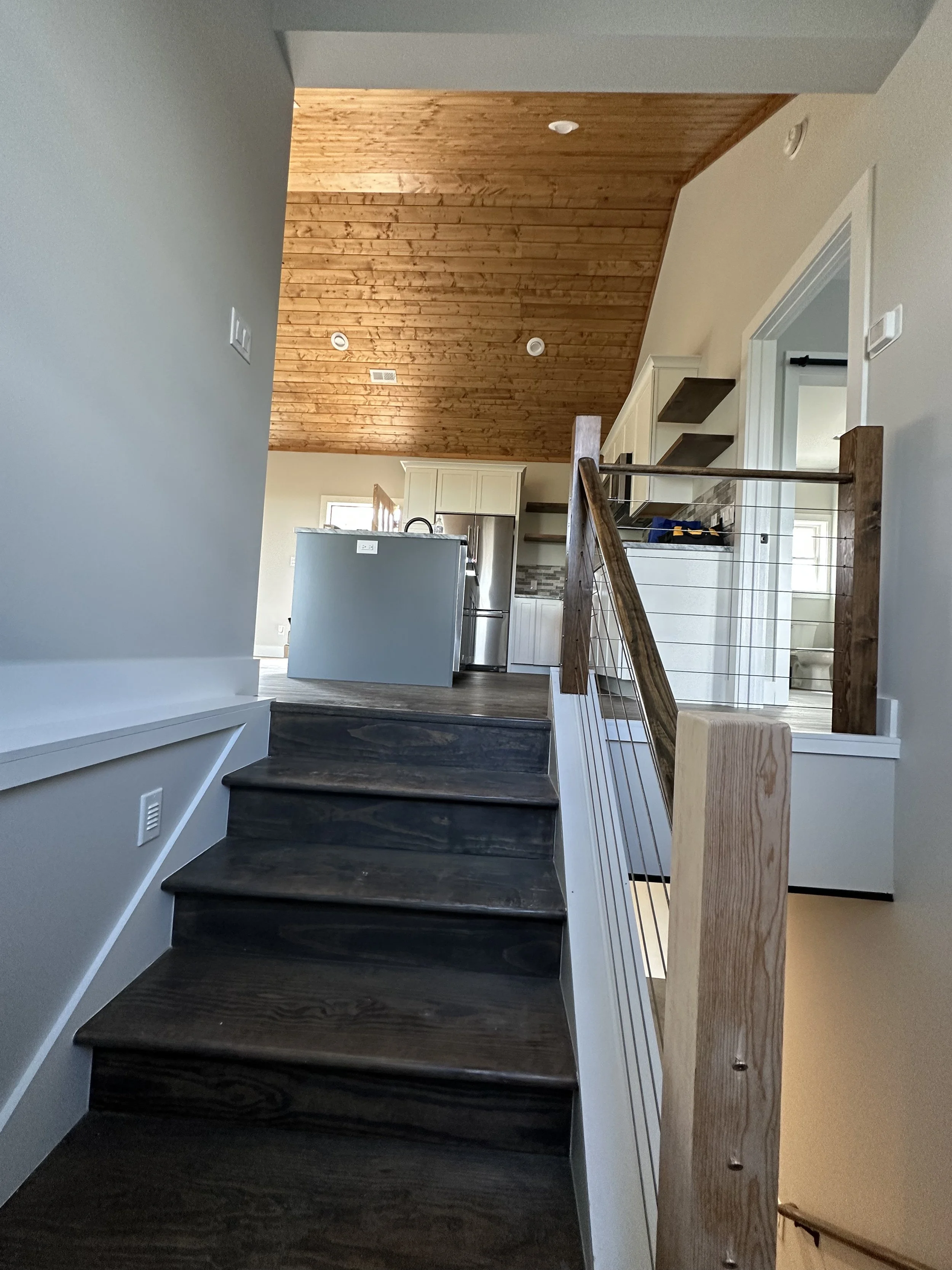 Interior view of a staircase with dark wooden steps, white walls, and a wooden handrail leading to a living area with a wooden ceiling, visible kitchen appliances, and a doorway.