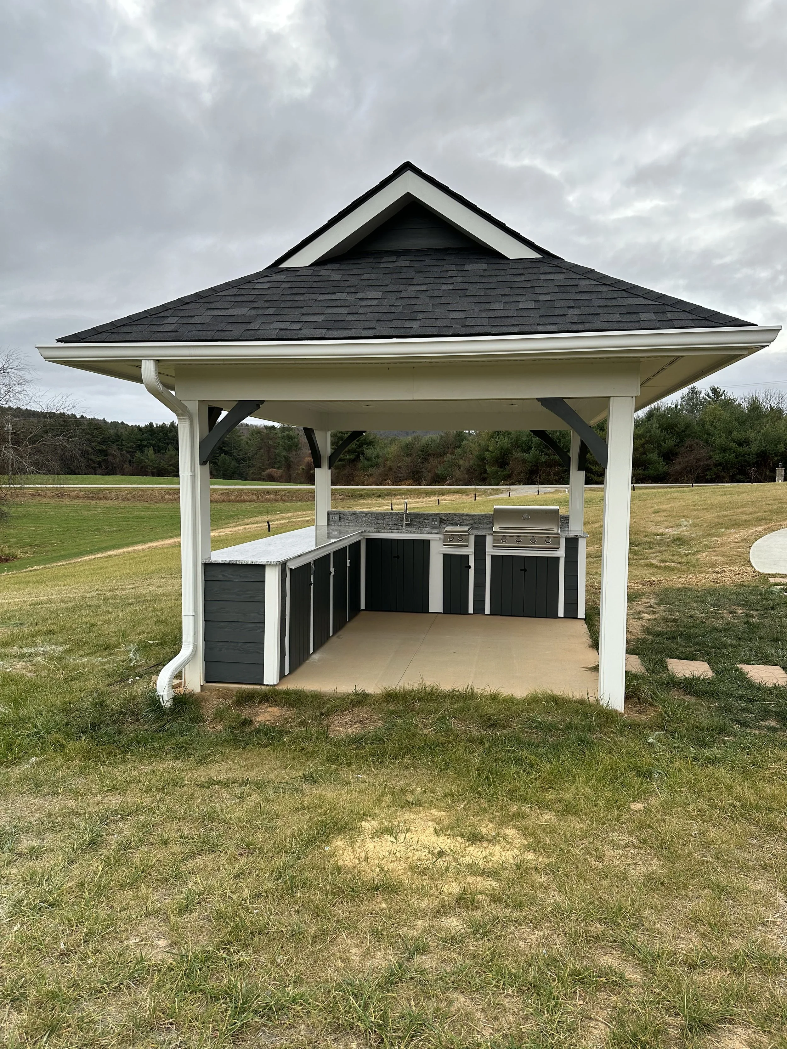 An outdoor gazebo with a built-in stainless steel grill and countertop, situated in a grassy field under a cloudy sky.