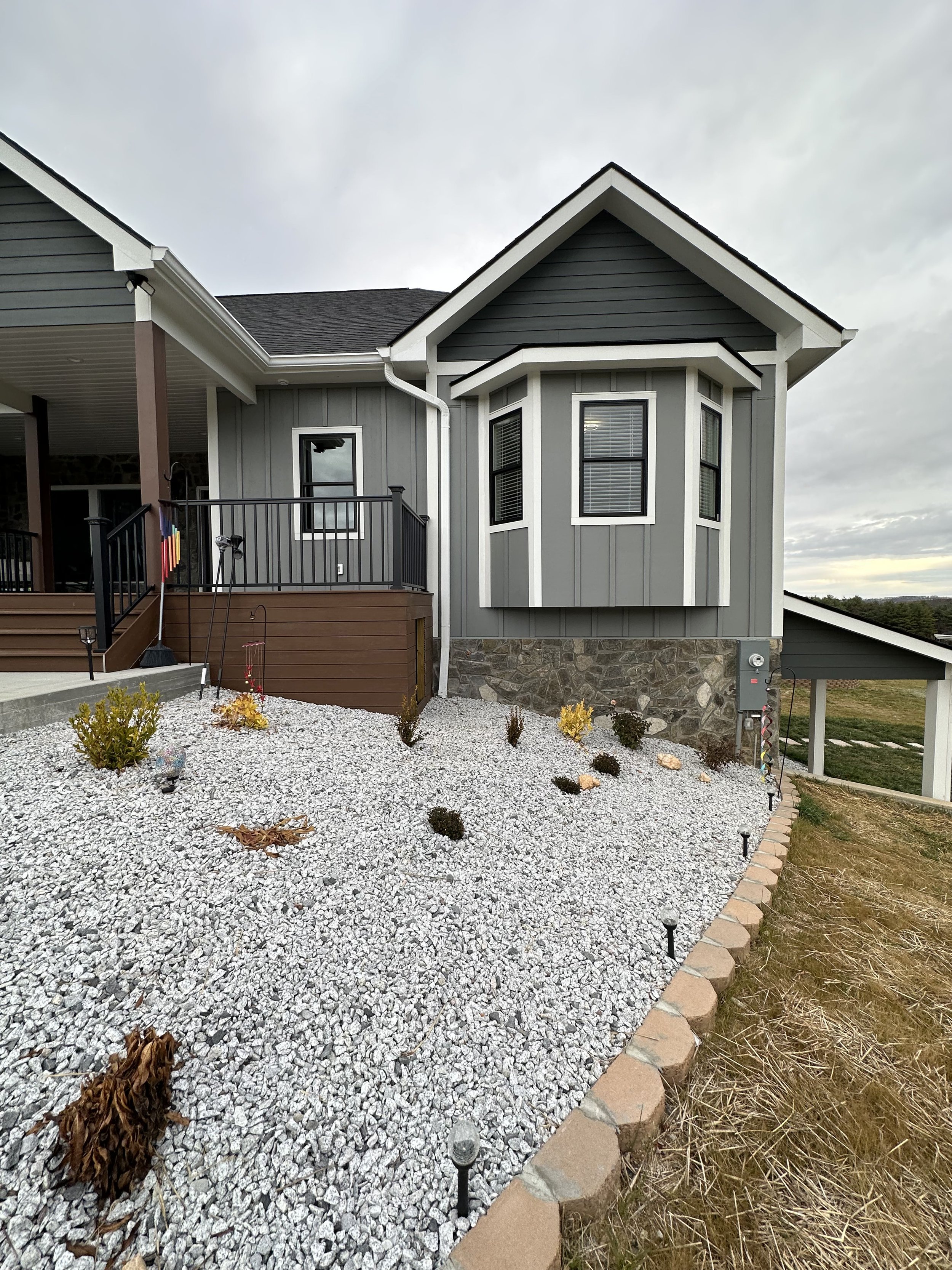 Front yard with white gravel and small bushes, bordered by bricks, next to a residential house with gray siding and windows, under a cloudy sky.