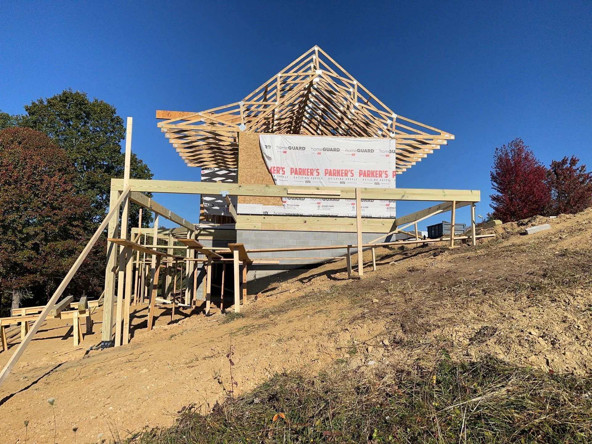 A house under construction with a wooden frame and roof trusses, surrounded by trees and a dirt ground, with a clear blue sky in the background.