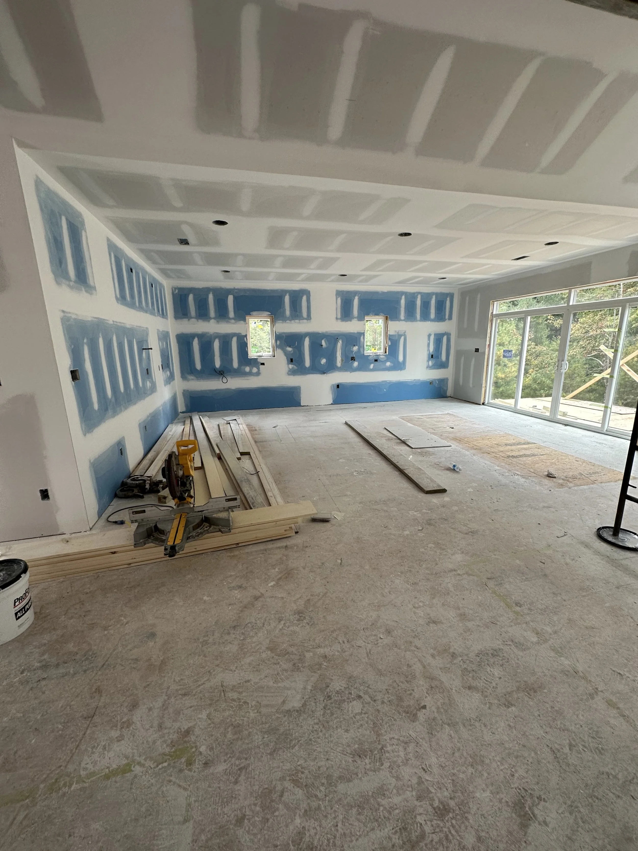 Living room under construction with drywall walls, unfinished ceiling, large sliding glass door, construction tools, wooden planks, and construction materials scattered on the floor.