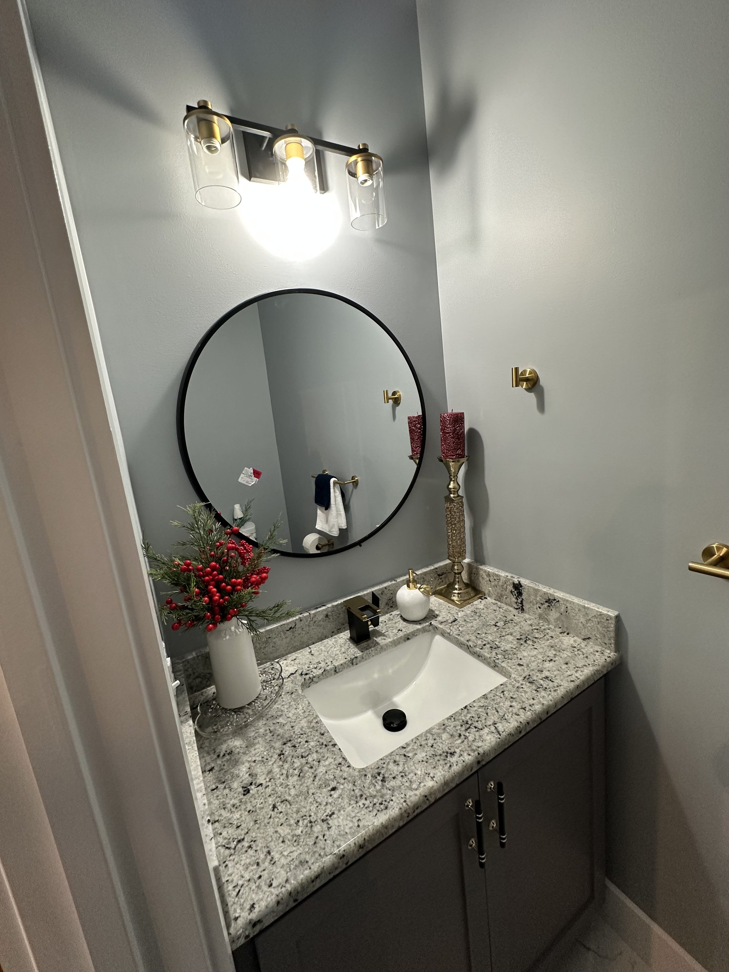 Bathroom vanity with a white sink, granite countertop, decorative candle holder, soap dispenser, mirror, and wall-mounted light fixture. A small Christmas tree with red berries and a spray of pine branches is on the counter.