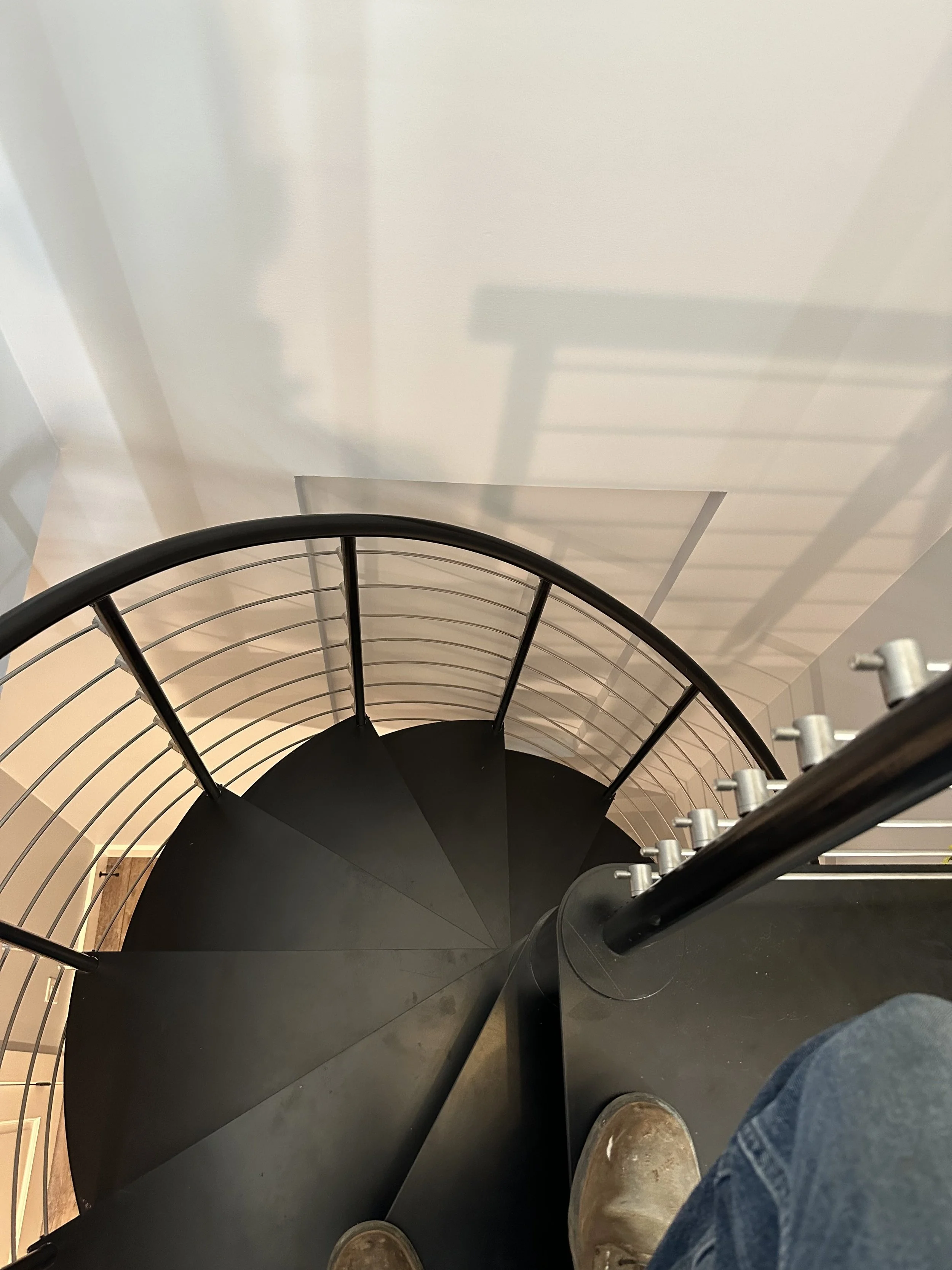 Top view of a black spiral staircase with a person wearing brown shoes and blue jeans standing at the bottom.
