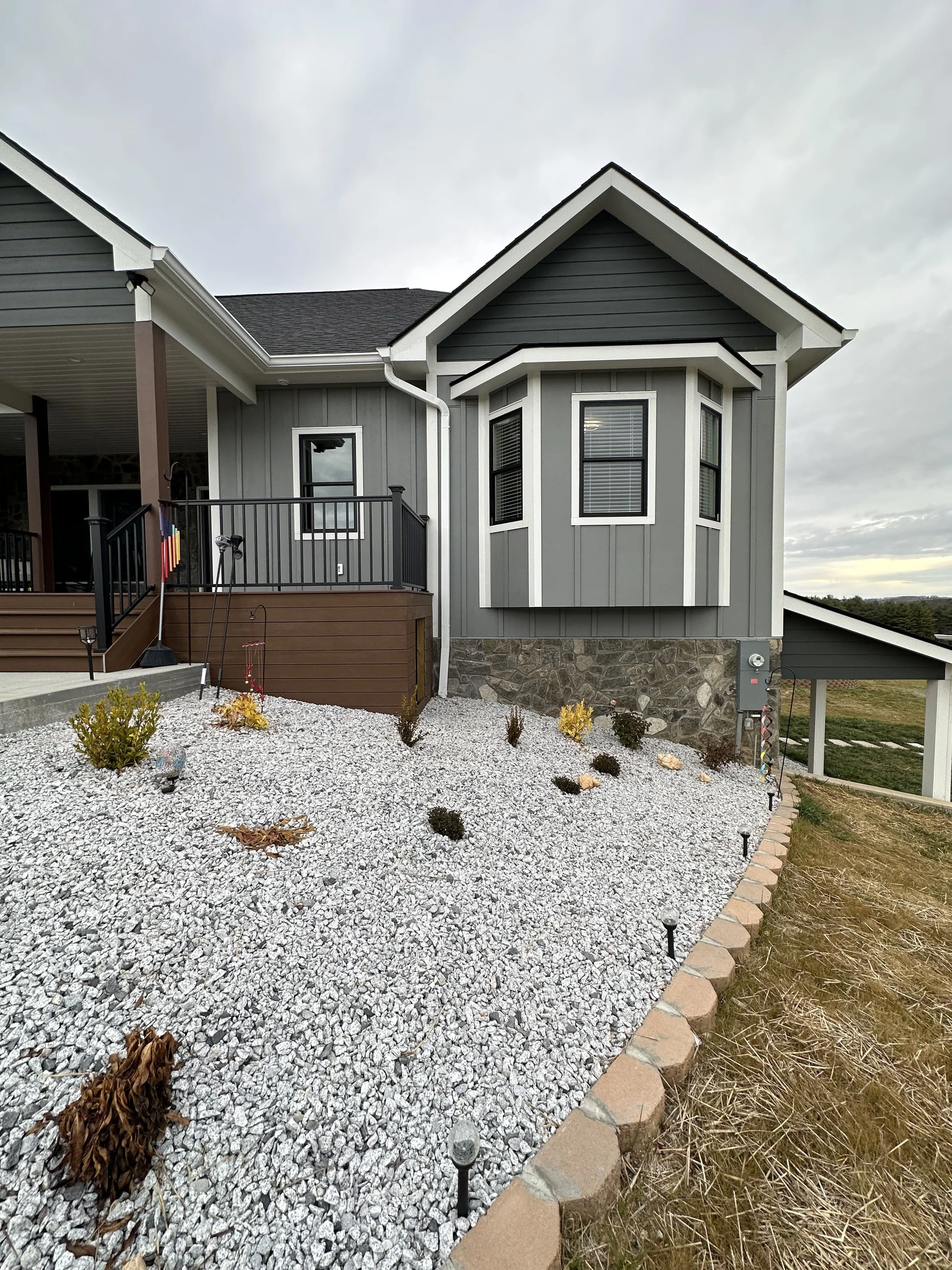 Front view of a house with gray vertical siding and a stone foundation, featuring a bay window, a small porch with black railing, a gravel yard with small bushes, and a lawn area to the right, under a cloudy sky.