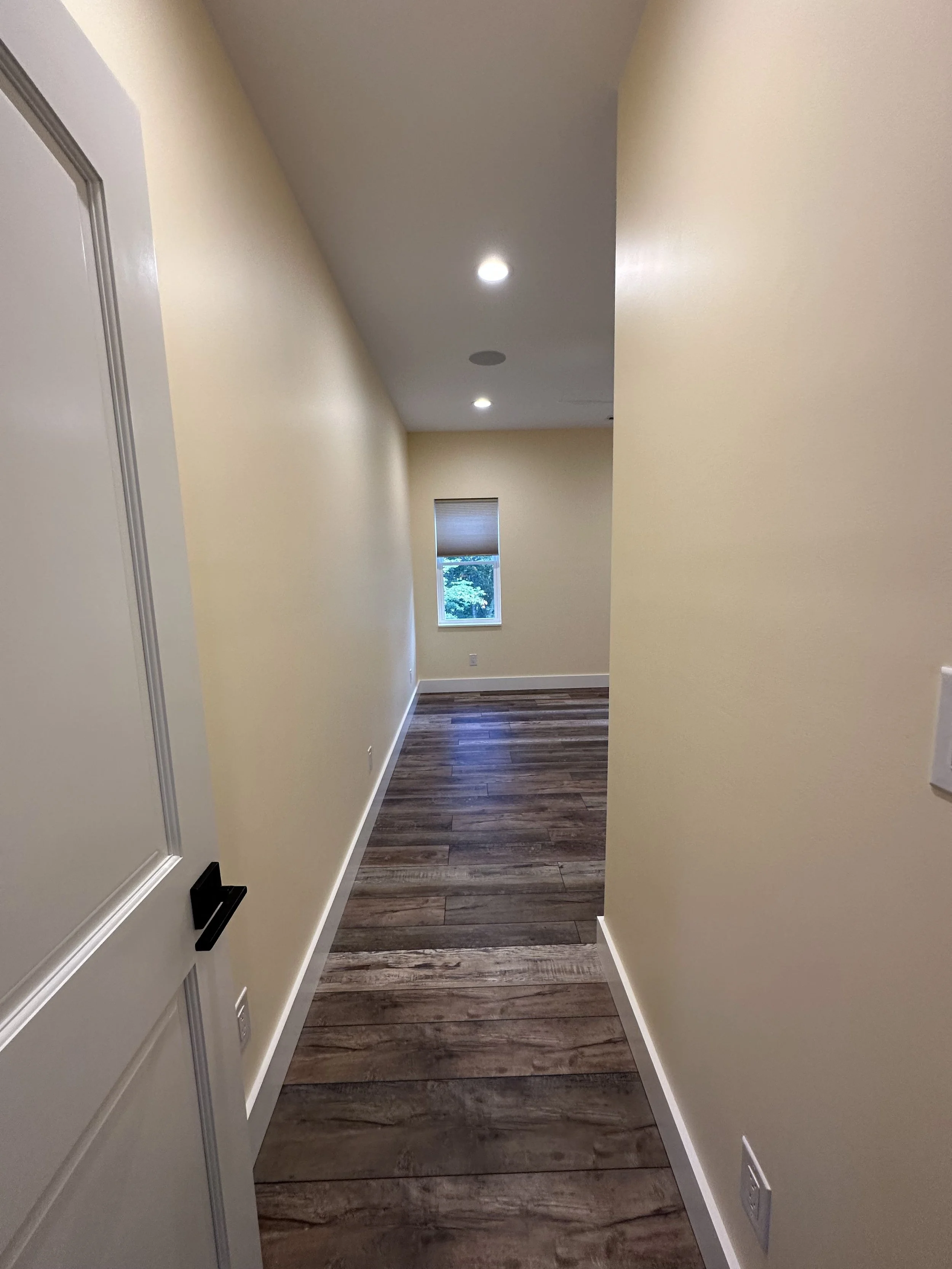 A narrow hallway in a house with beige walls, a wooden floor, a window at the end, and ceiling lights.