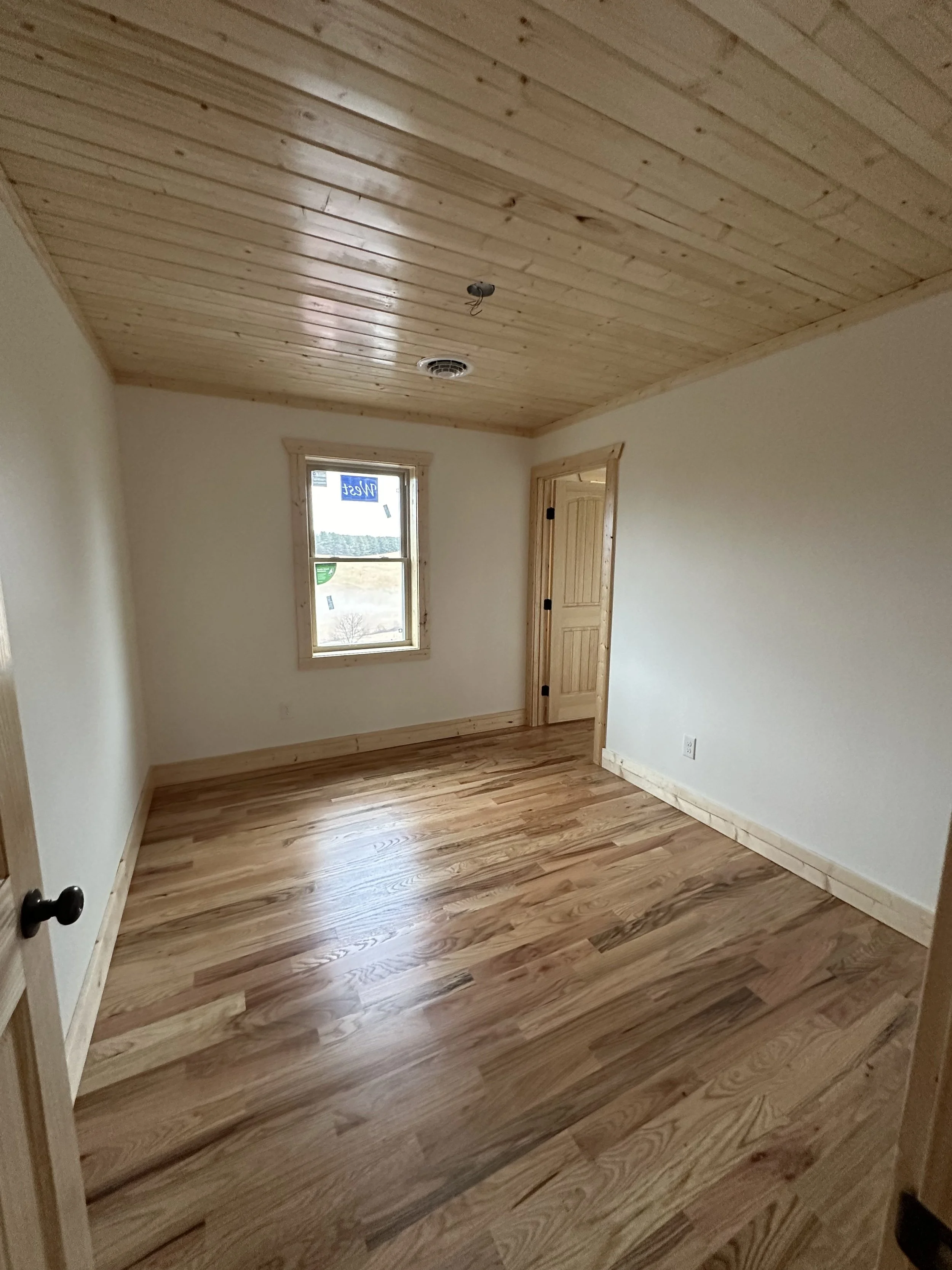 Empty room with wooden floor, wooden ceiling, and a window showing a landscape outside, including hills and trees.