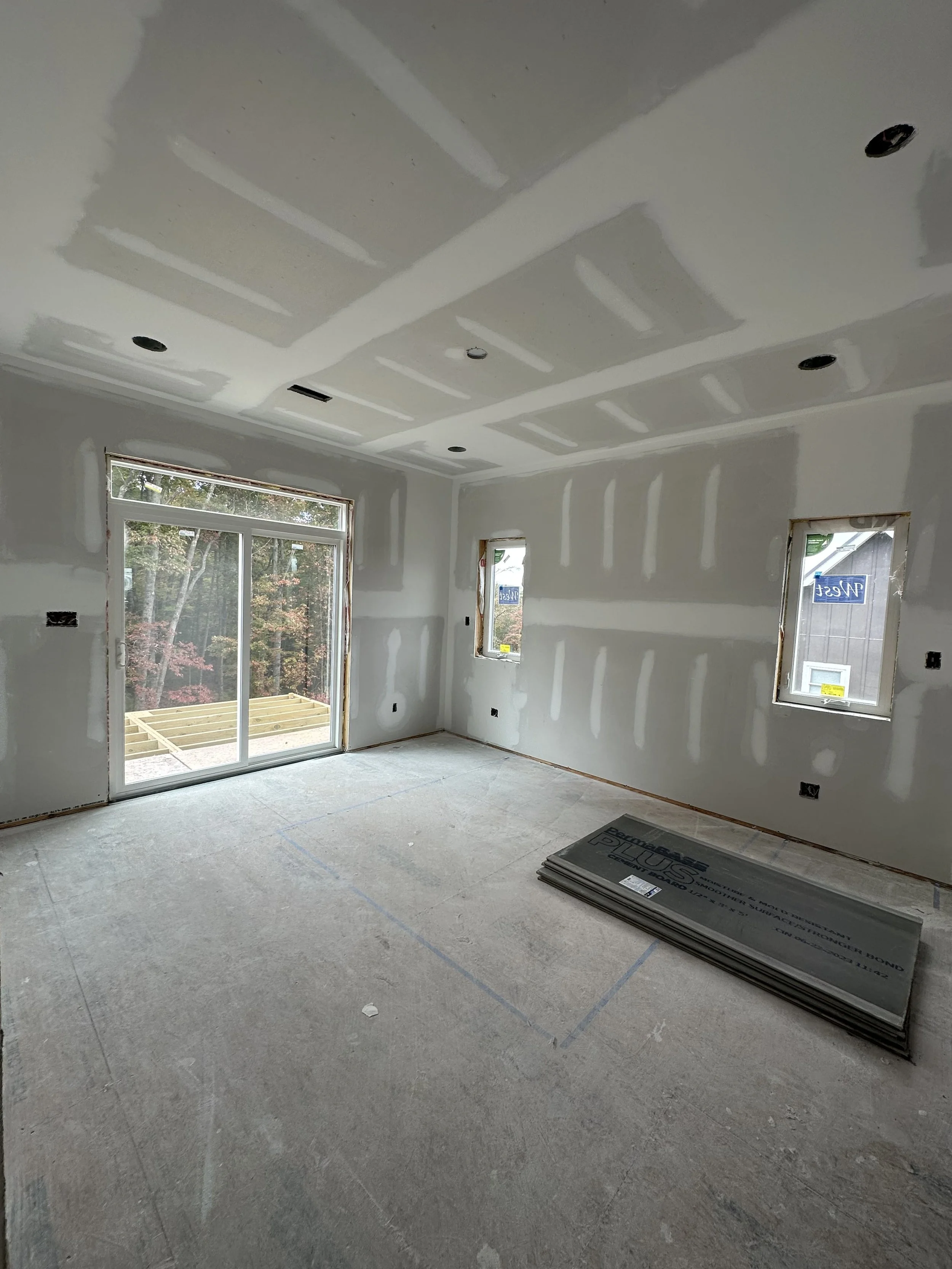 An unfinished room with drywall installed and seams taped, ready for painting. The room has large windows, including a sliding glass door leading to an outdoor area with a wooden deck. Construction materials are stacked on the floor.