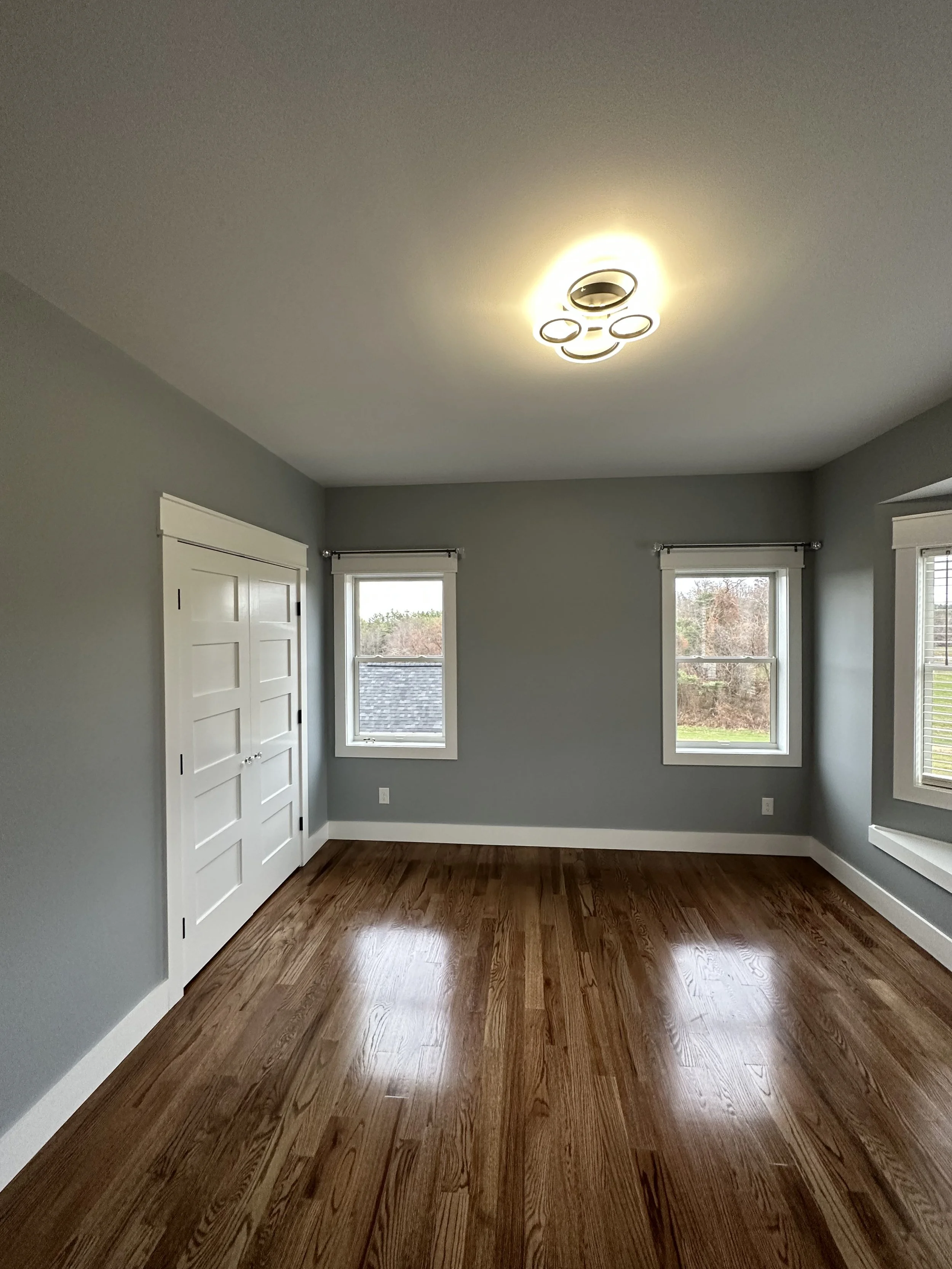 Empty room with hardwood floors, gray walls, white trim, and three windows with white blinds, showing an outdoor view of trees and a cloudy sky.