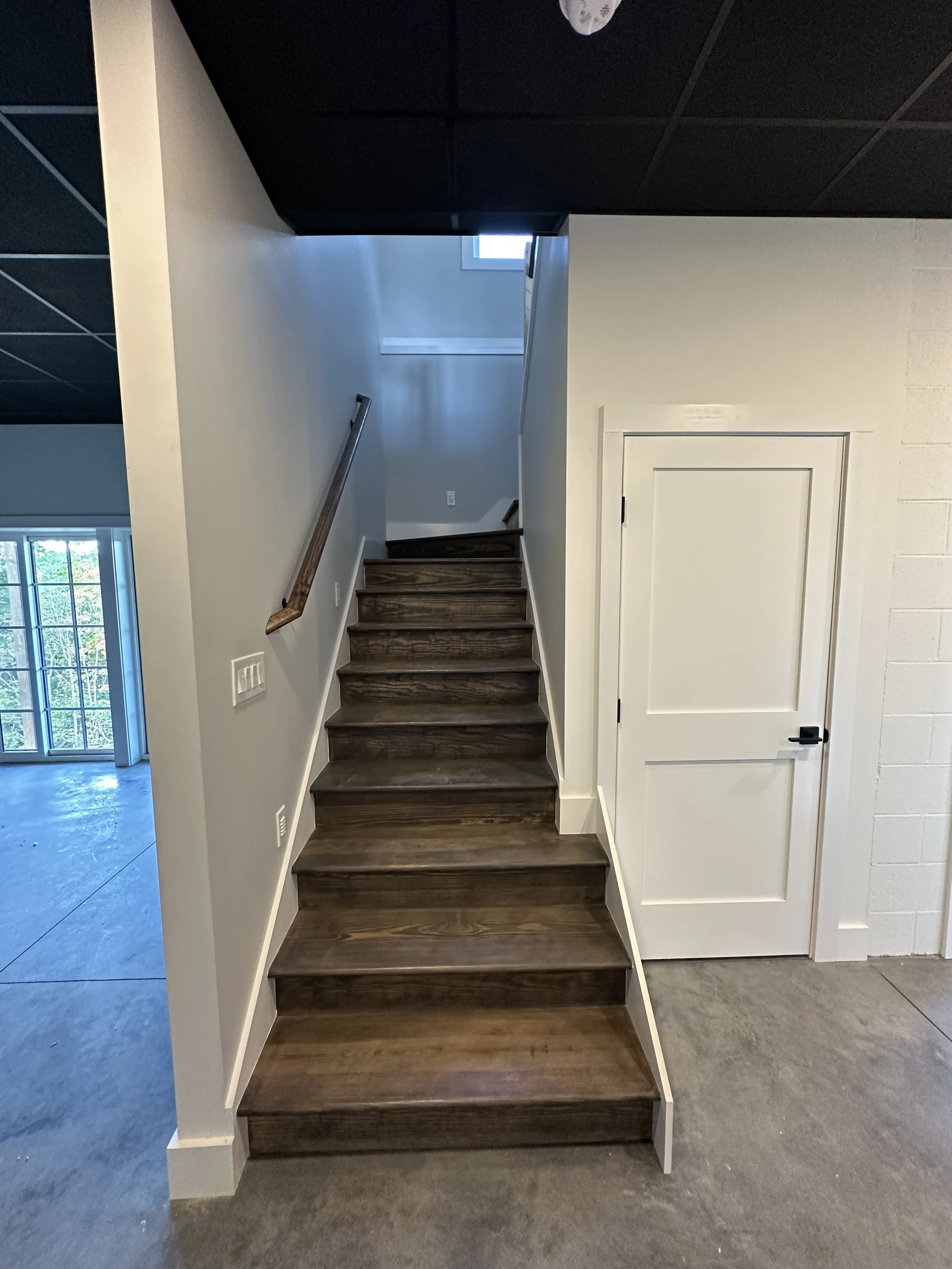 Wooden staircase with dark brown steps leading upward in a modern interior space, with a wooden handrail on the left side. To the right, a white door with black hardware is visible. The walls are painted white, and a small window at the top of the st