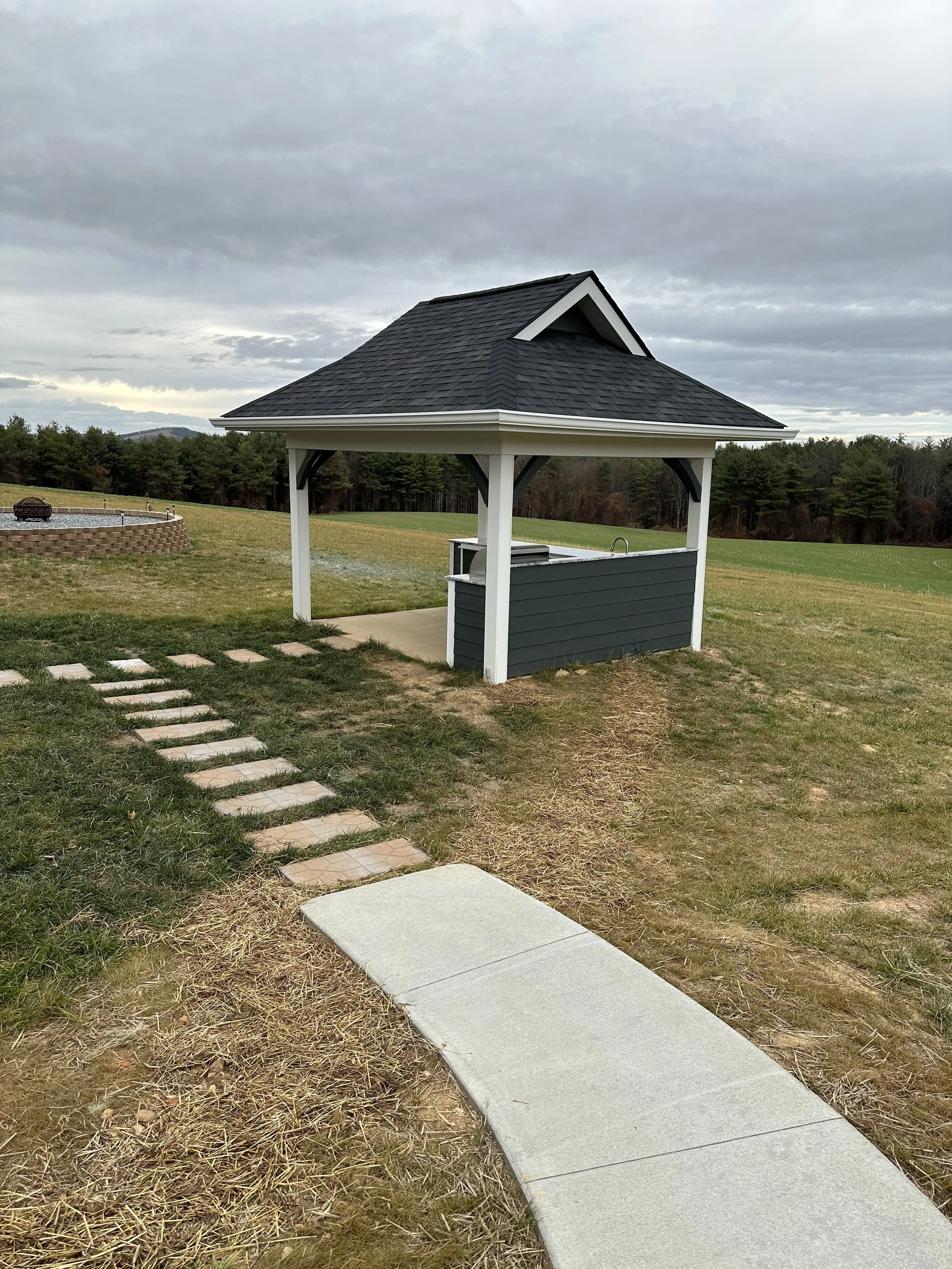 An outdoor scene with a gazebo that has a dark shingled roof and white supporting columns, a gray countertop with a faucet, a stone pathway, and a grassy area with a fire pit and trees in the background, under a cloudy sky.