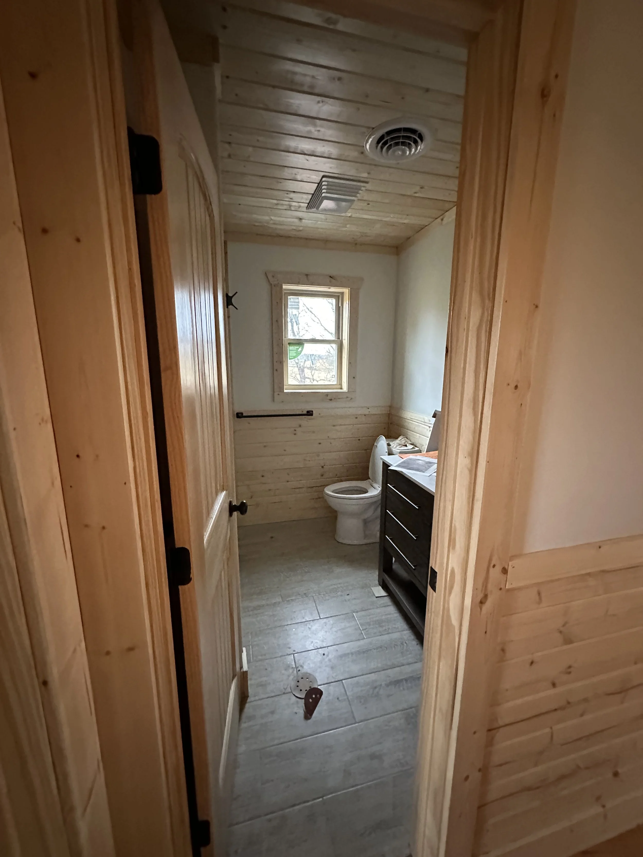 A bathroom with wooden walls and ceiling, a small window, a toilet, a dark-colored vanity, and a few scattered objects on the floor.