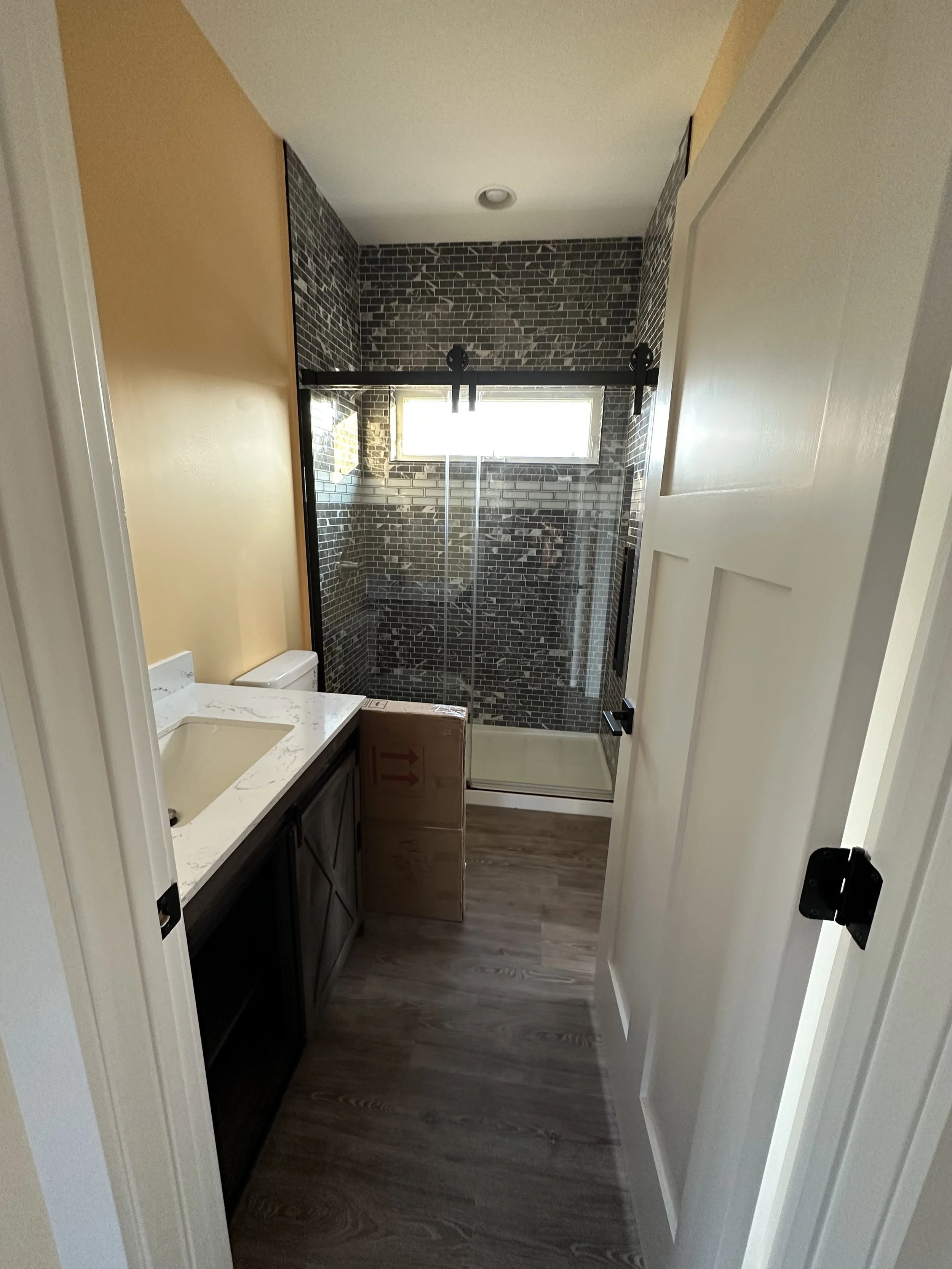 A bathroom with a white door, dark vanity with a marble countertop, partially visible toilet, and a glass-enclosed shower with black-framed sliding doors and brick-patterned black and gray tiles, a small window.