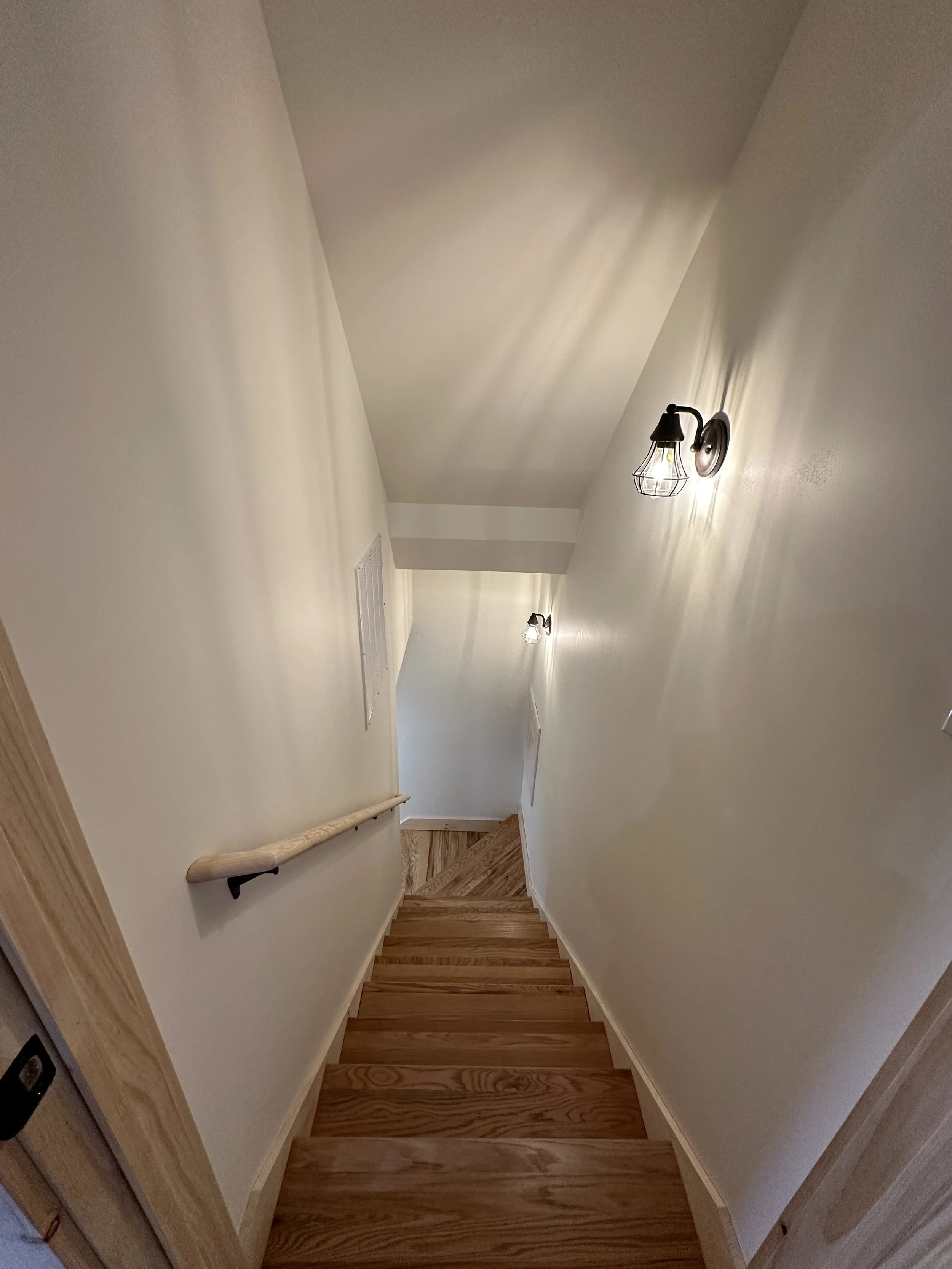 Wooden staircase with beige walls, a handrail on the left side, wall-mounted light fixtures, and wooden flooring at the bottom.