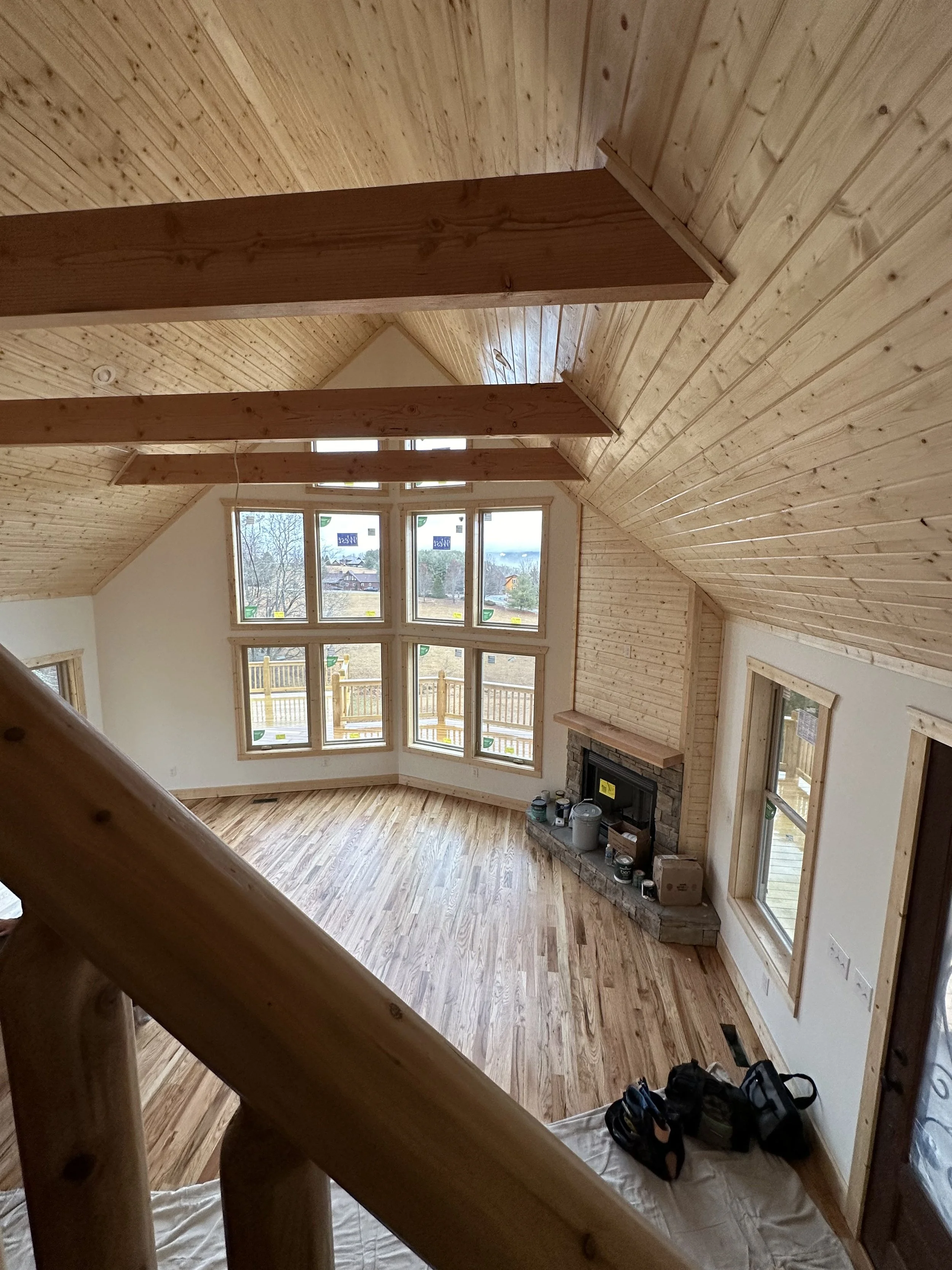 Interior view of a house with wooden ceiling and floor, large windows showing an outdoor landscape with trees and houses, a stone fireplace, and some construction tools on the floor.