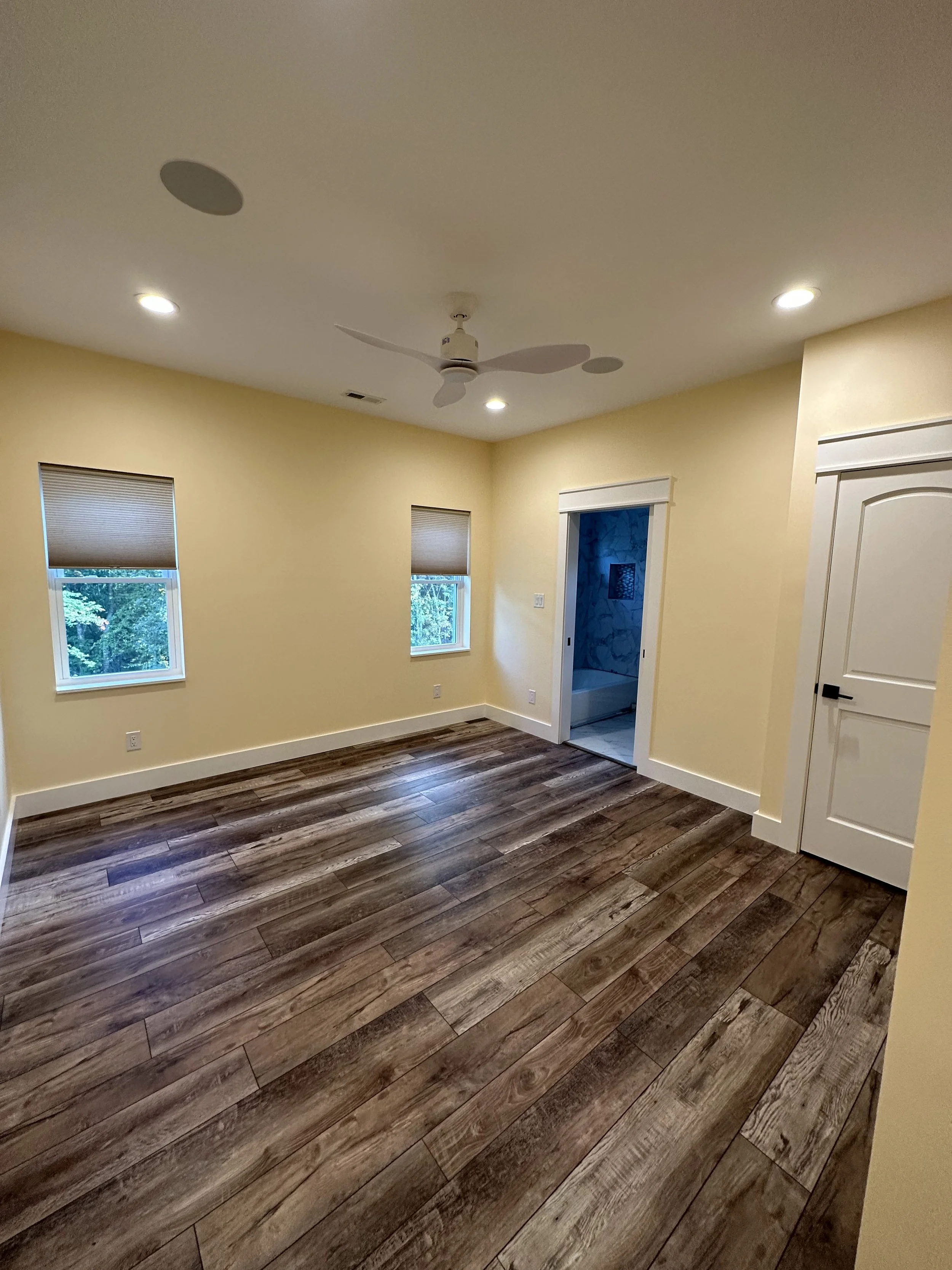 Empty room with hardwood flooring, beige walls, two windows with blinds, a ceiling fan, and a doorway to a bathroom with marble tiles.