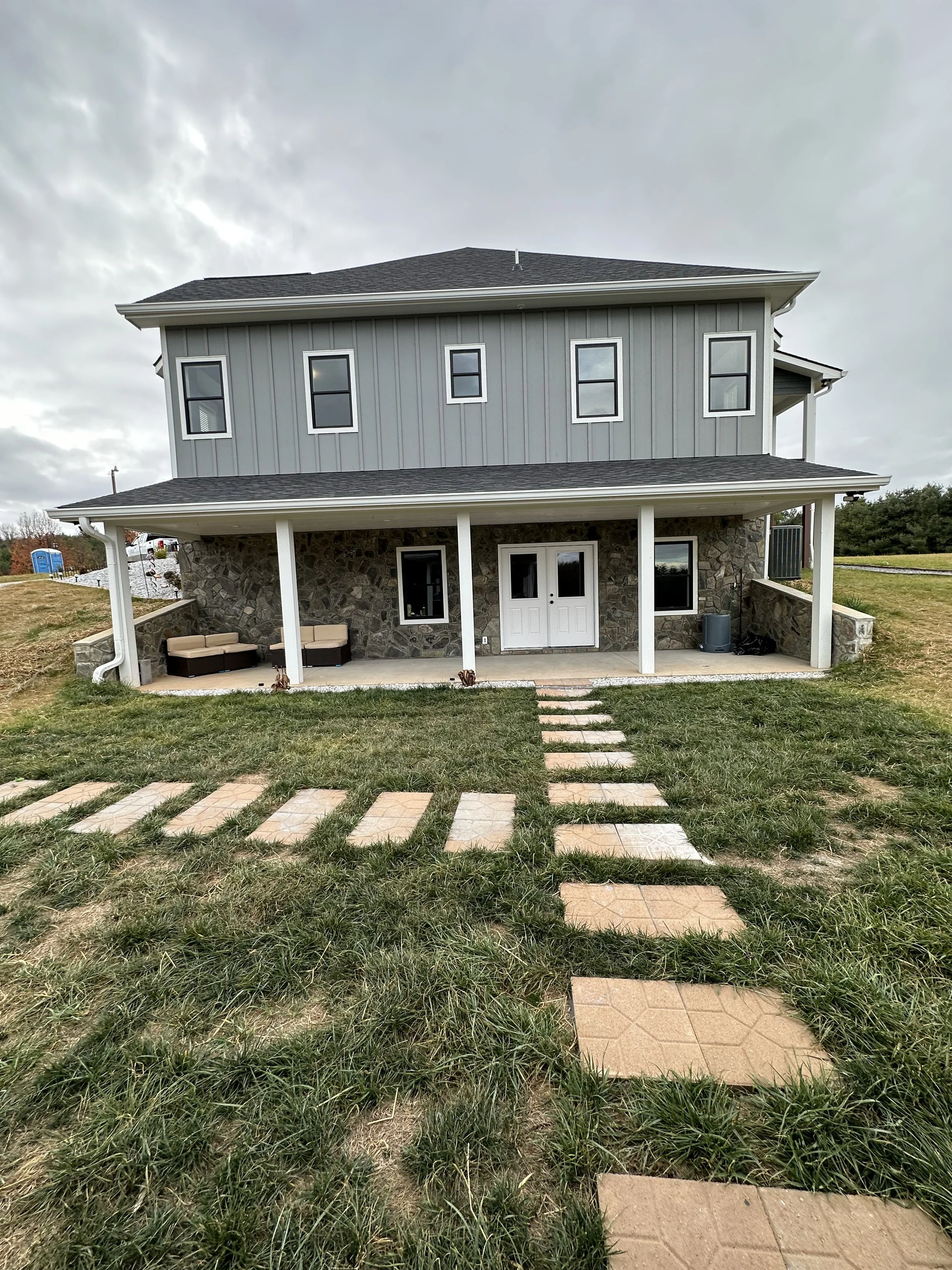 A two-story house with a stone and gray siding exterior, featuring a covered porch with pillars, a white front door, and several windows. The front yard has a pathway made of stepping stones leading to the house and is surrounded by grass. The sky is