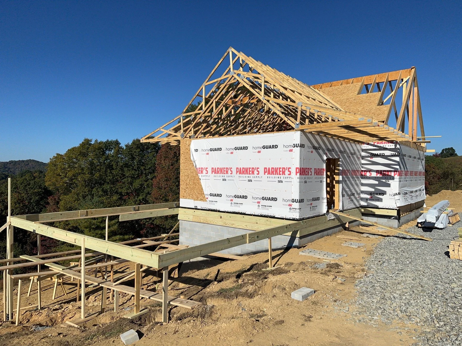 A house under construction with wooden framing, insulation wrapped in plastic, and a clear blue sky in the background.