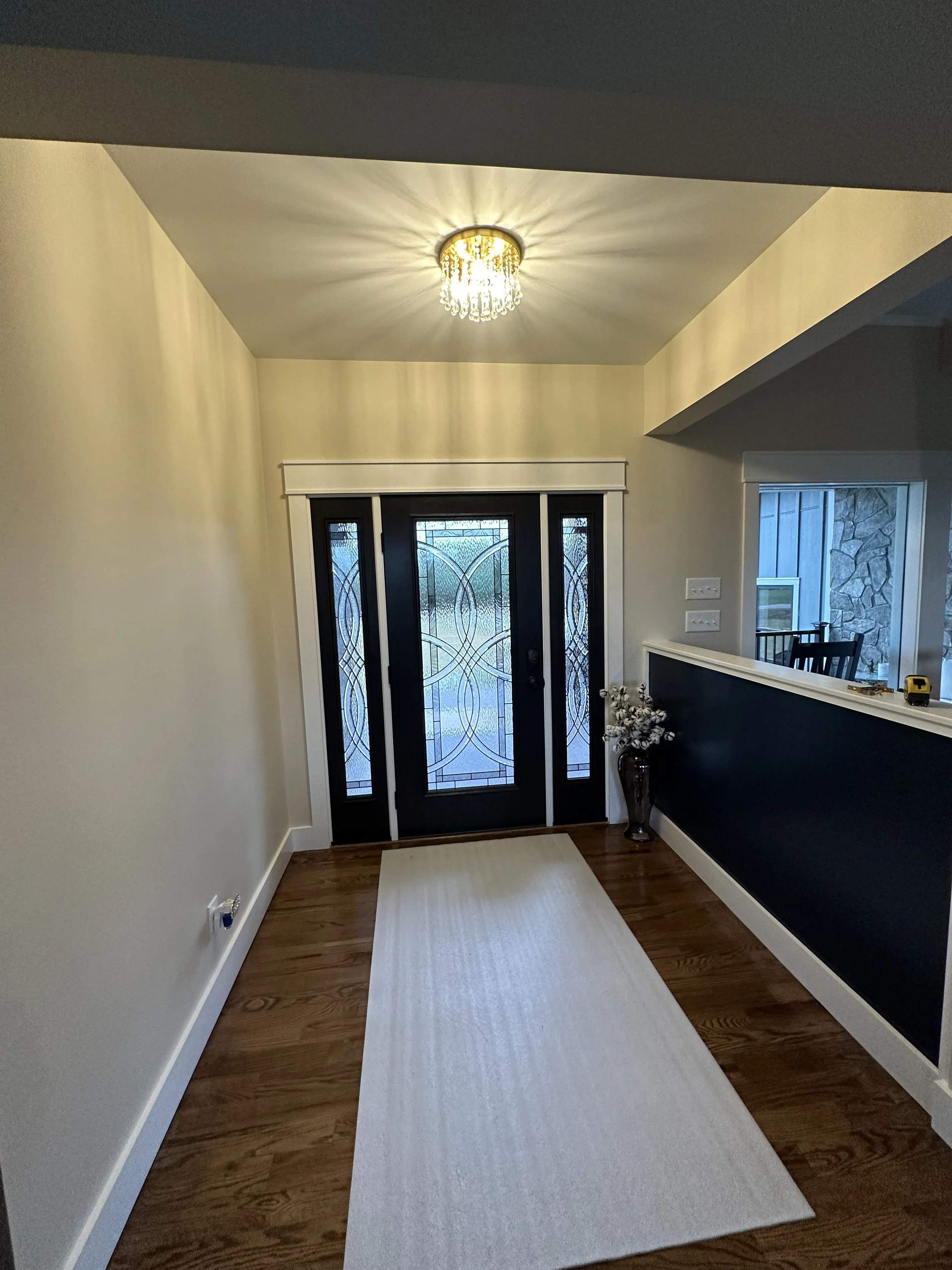 A front entryway with a black door featuring decorative glass panels, a cream-colored runner rug on wooden flooring, a vase with flowers in the corner, and a chandelier light fixture on the ceiling.