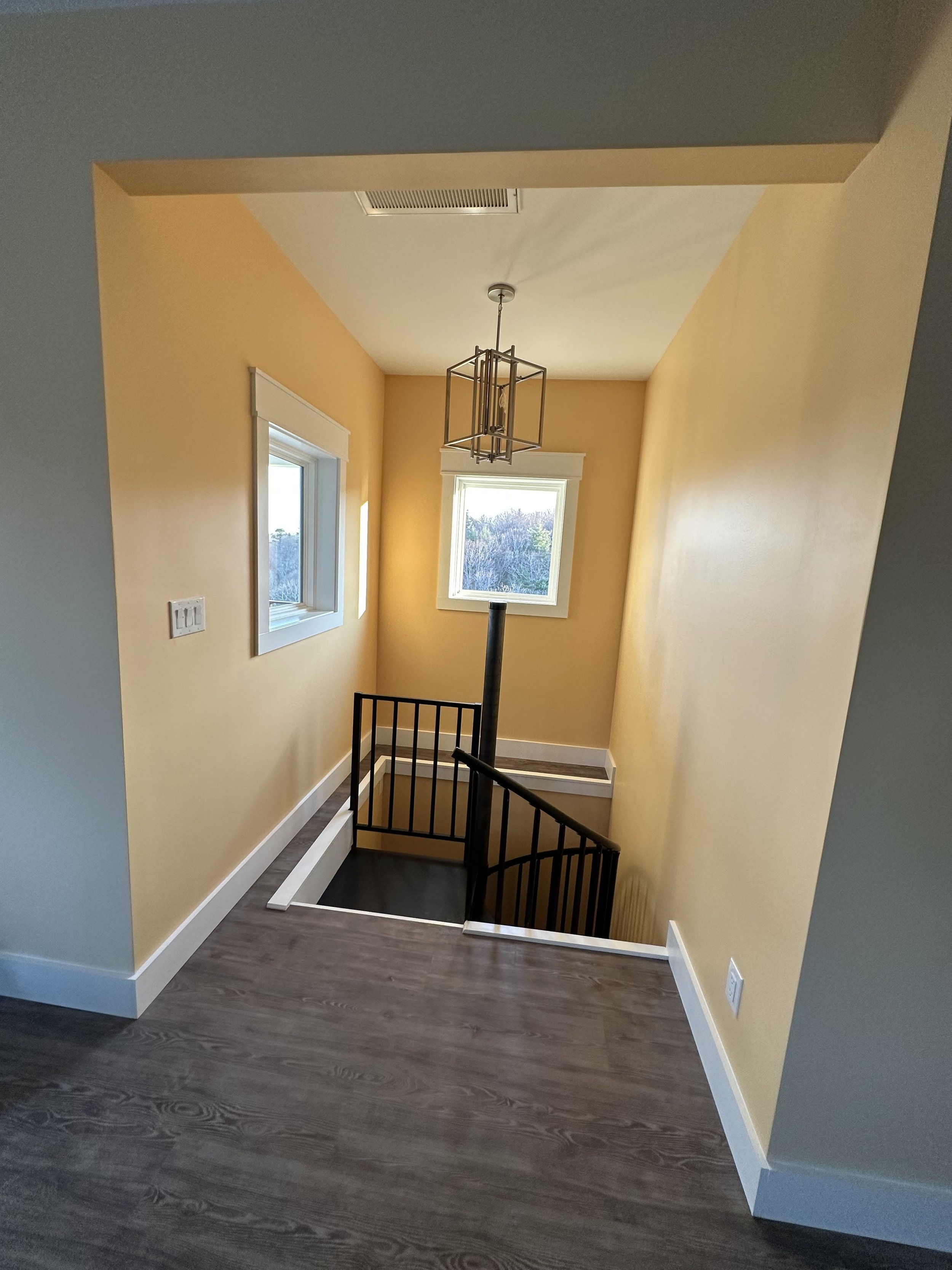 Interior view of a stairwell with yellow walls, two windows, a hanging light fixture, and dark wood flooring.
