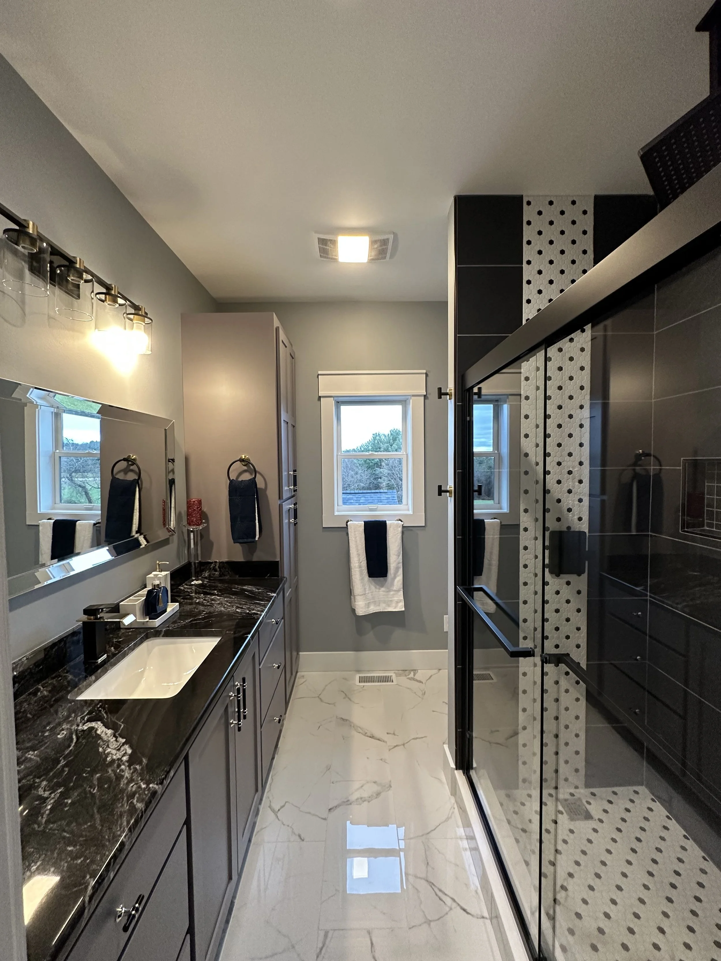 A modern bathroom with a black marble countertop vanity with a white sink, a large mirror, and a window above it. To the right, a black glass shower enclosure with geometric black and white tile accent wall. Light gray walls, marble floor, and natural light from two windows.
