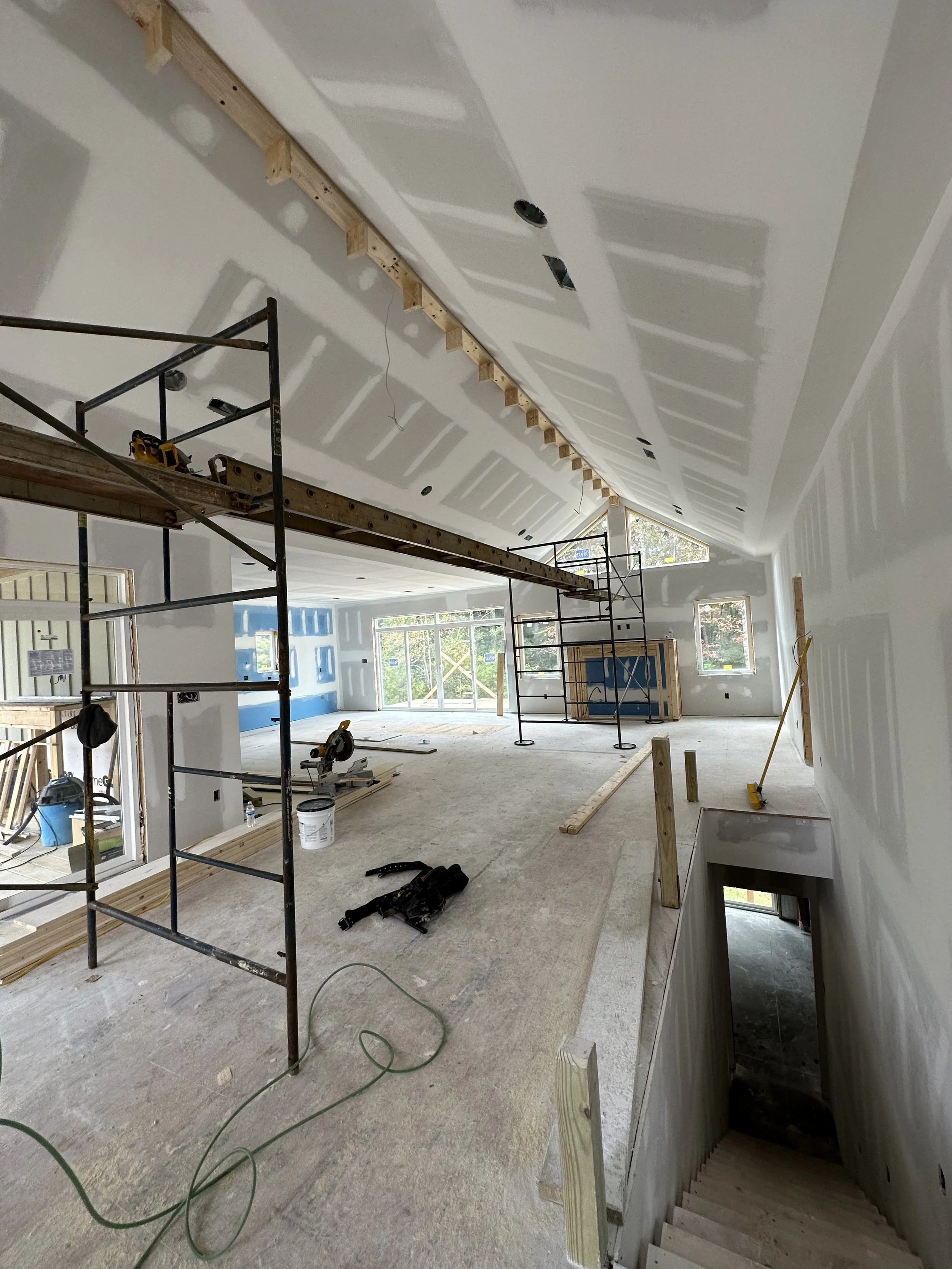 Interior of a building under construction with scaffolding, unfinished walls, and construction tools. The ceiling has drywall installed with some cutouts for lighting fixtures and vents. Large windows provide natural light.