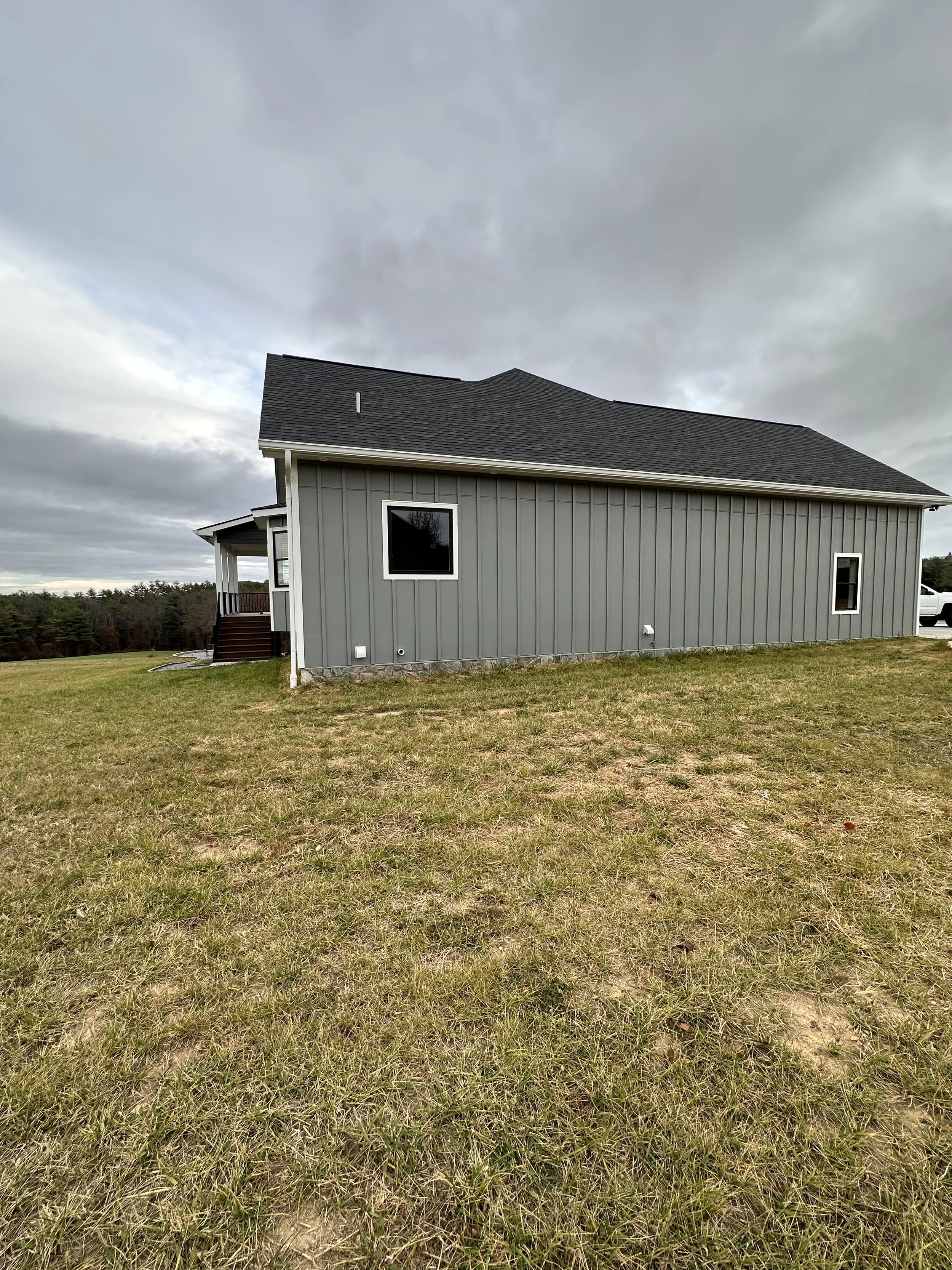 A house with gray siding and a dark gray roof, situated on a large grassy lawn with a cloudy sky overhead.