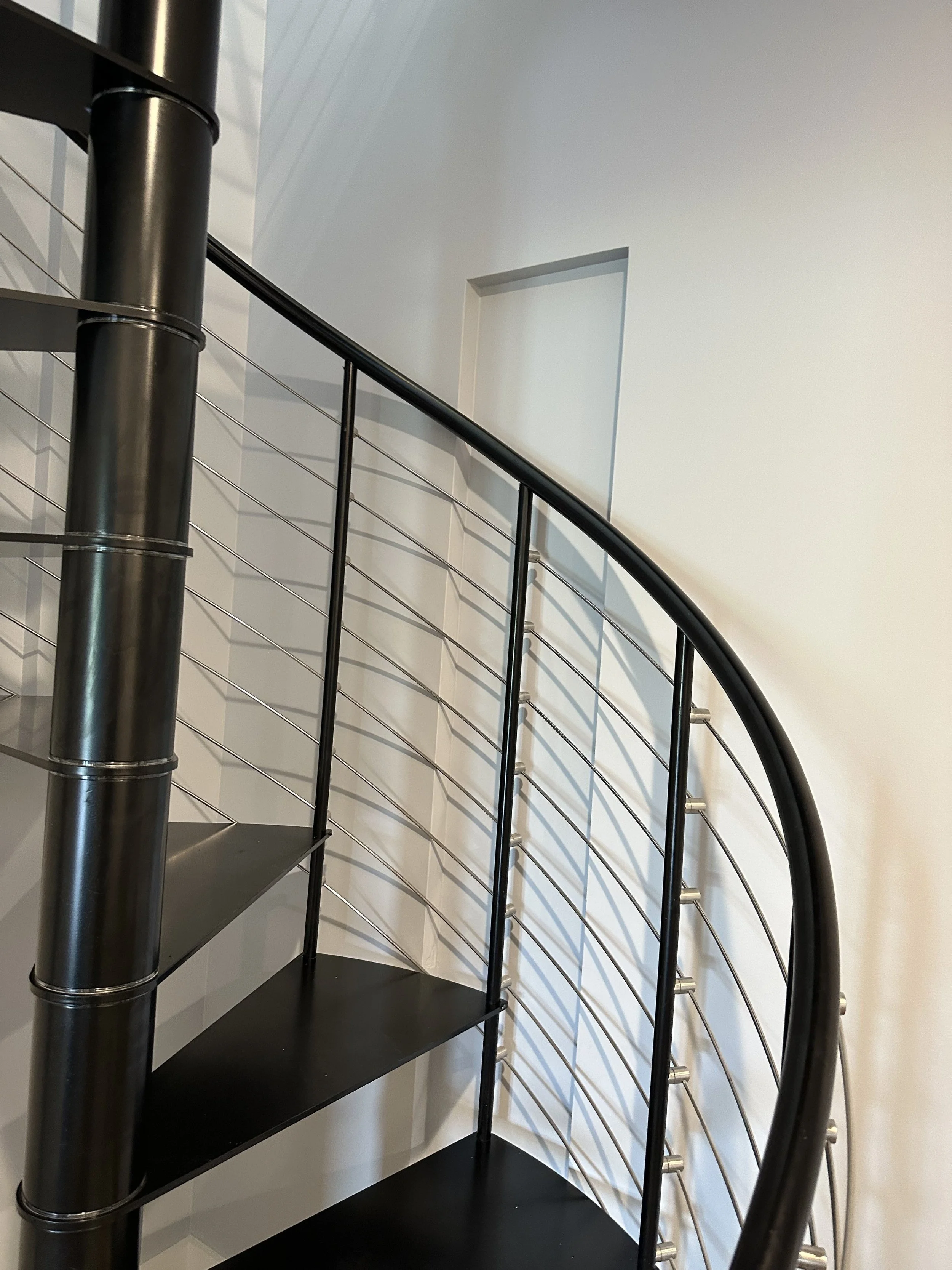 A spiral staircase with black steps, metallic railing, and white wall background. A rectangular opening in the wall visible at the top of the staircase.