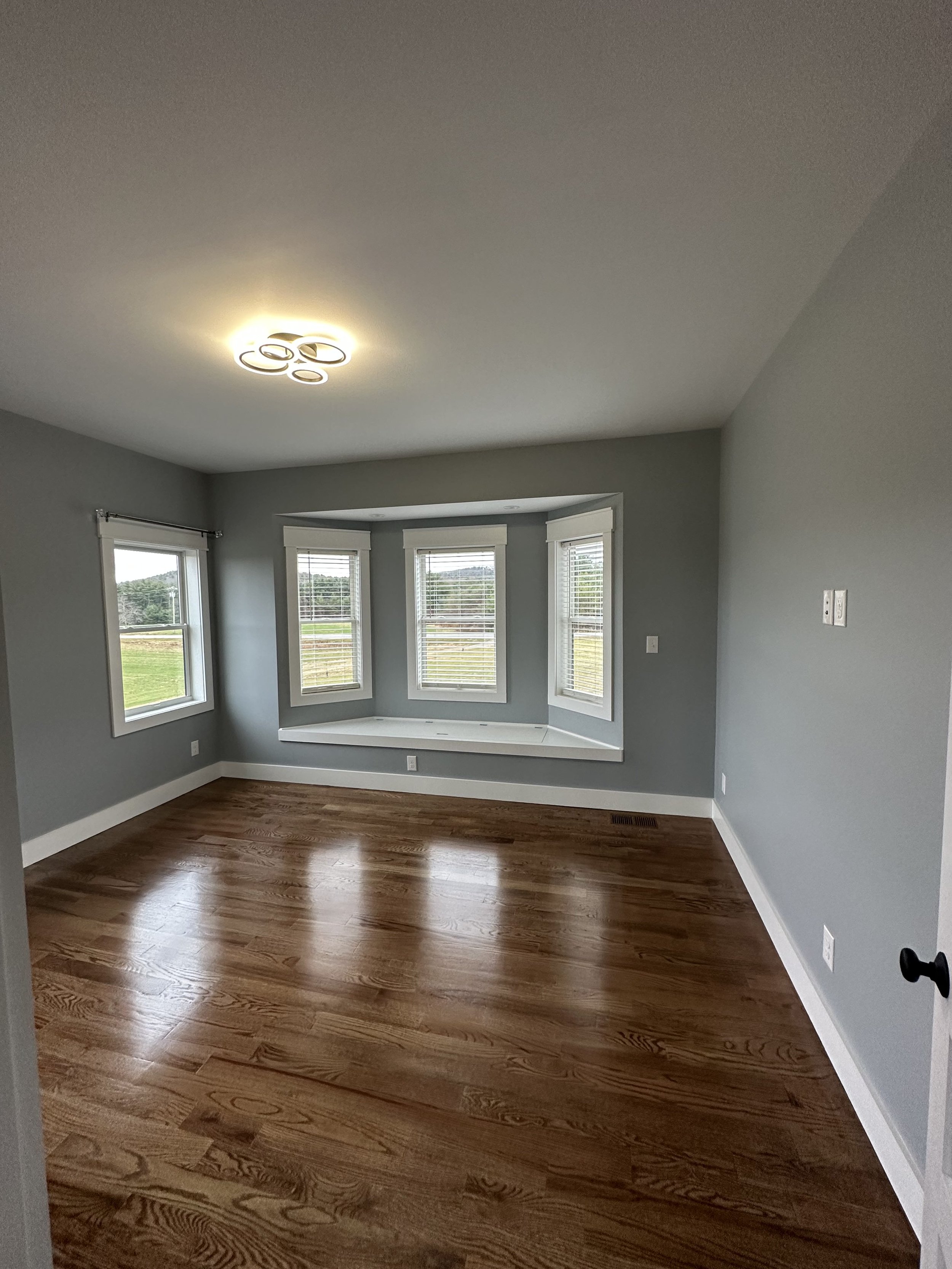 Empty room with three large windows, hardwood floors, gray walls, white trim, and a ceiling light fixture.