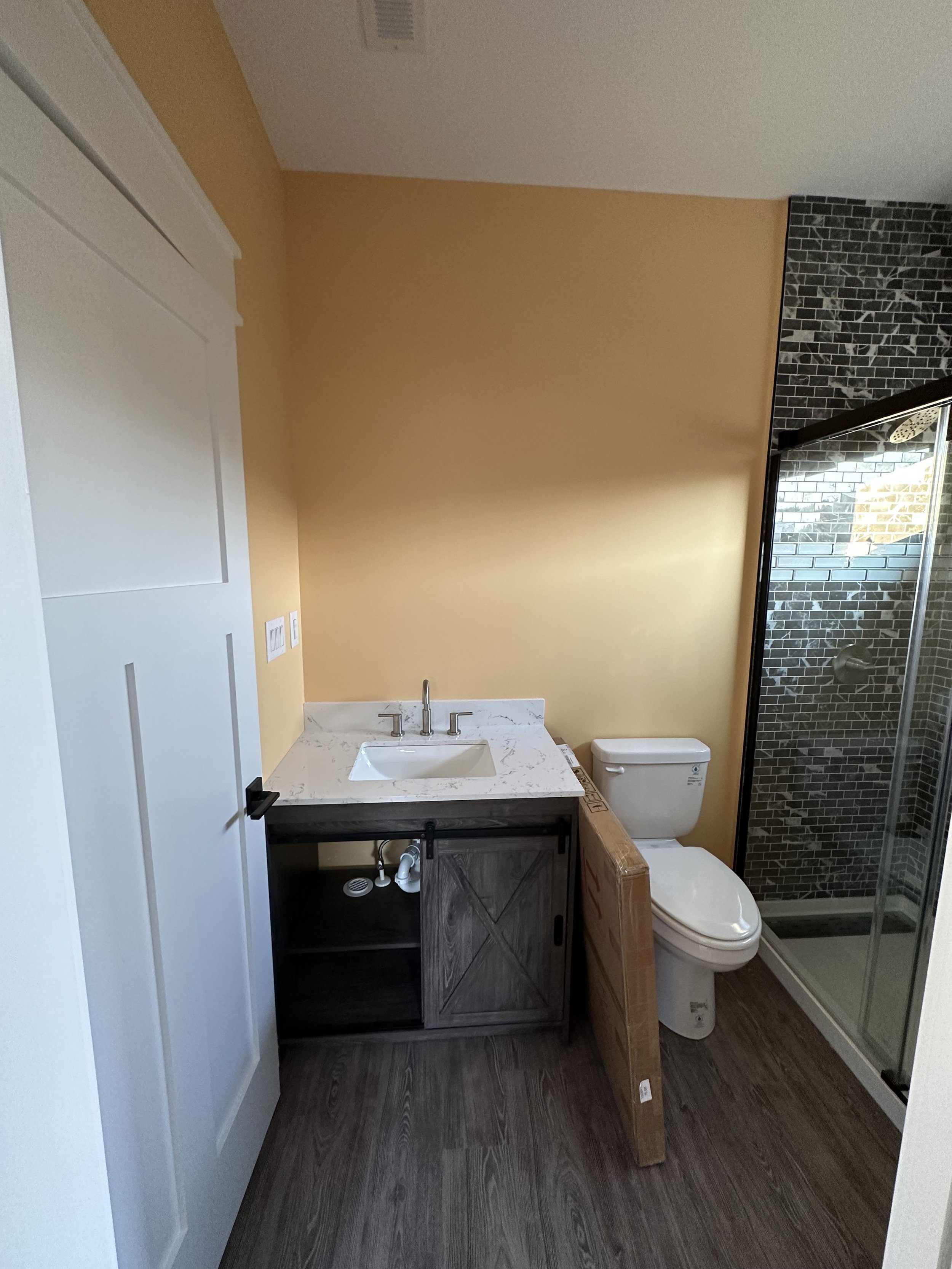 A bathroom with a yellow accent wall, a white ceiling with a vent, a dark wood vanity with a white marble countertop, a modern sink with a faucet, an open cabinet underneath, a toilet, and a shower with dark subway tile and a glass door.