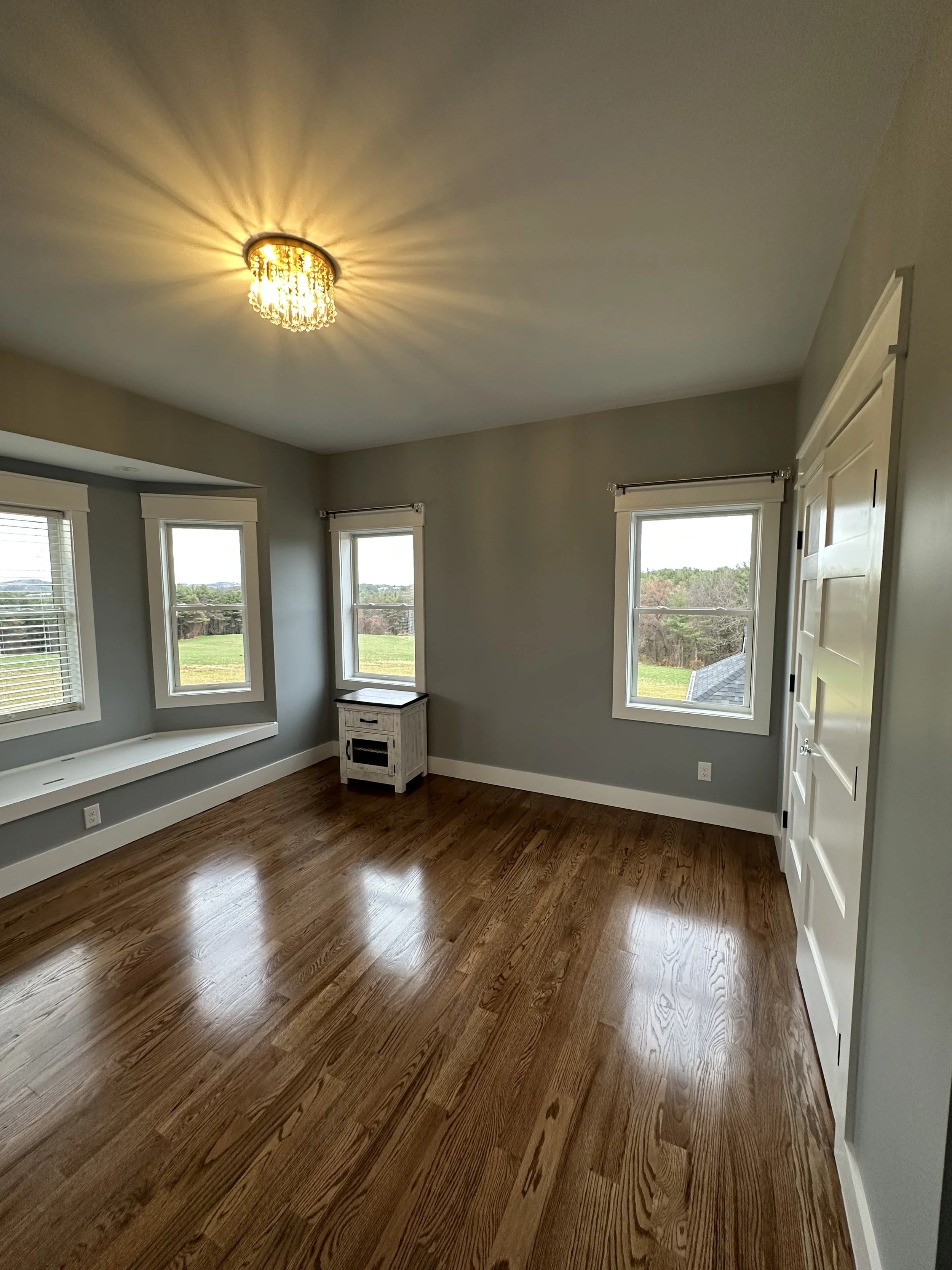 A room with hardwood floors, gray walls, large windows showing a green outdoor landscape, a white window seat, and a ceiling light with a decorative crystal fixture.