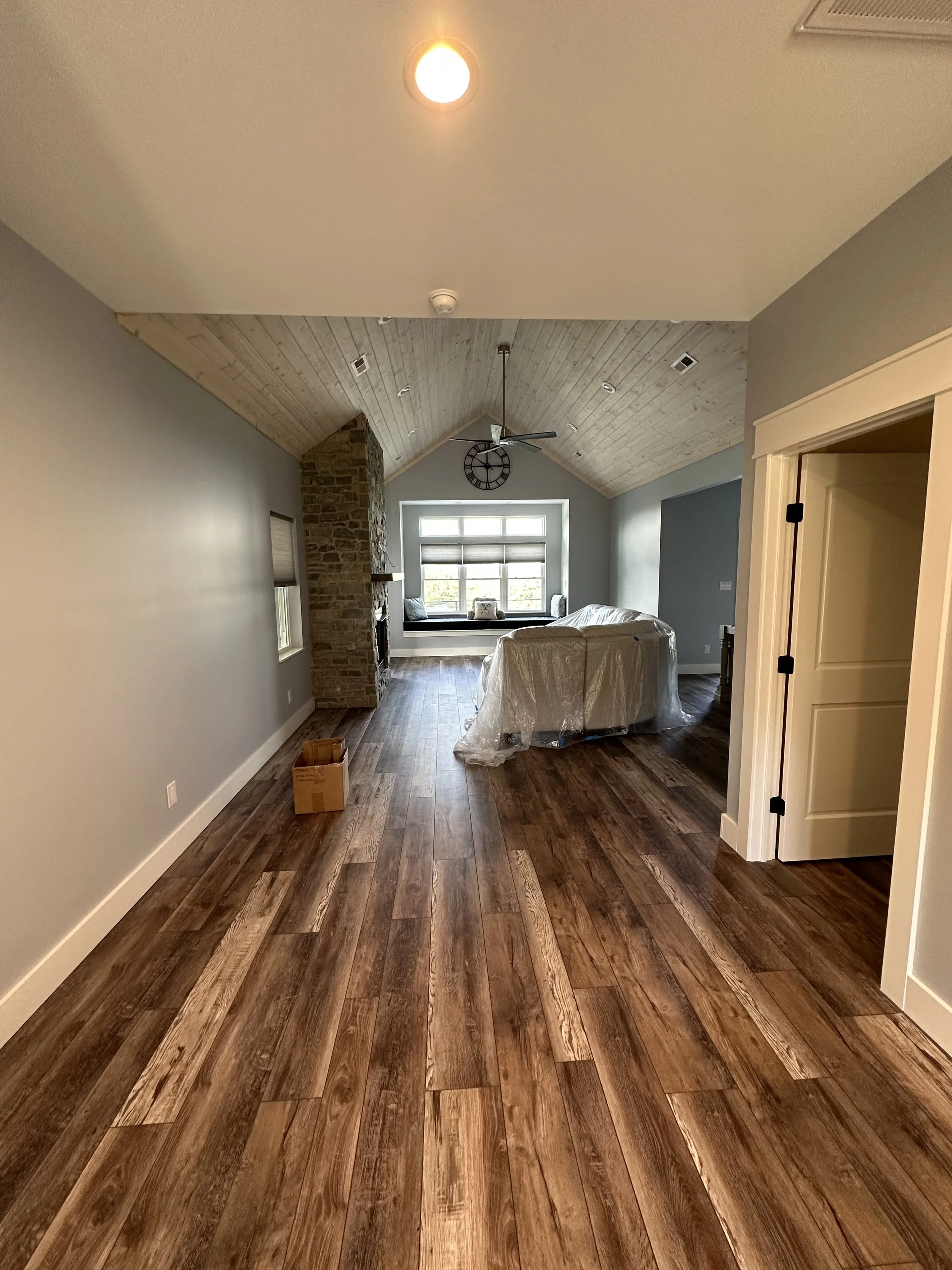 Living room with wood floors, a large window seat, stone fireplace, ceiling fan, and a vaulted ceiling with recessed lighting.