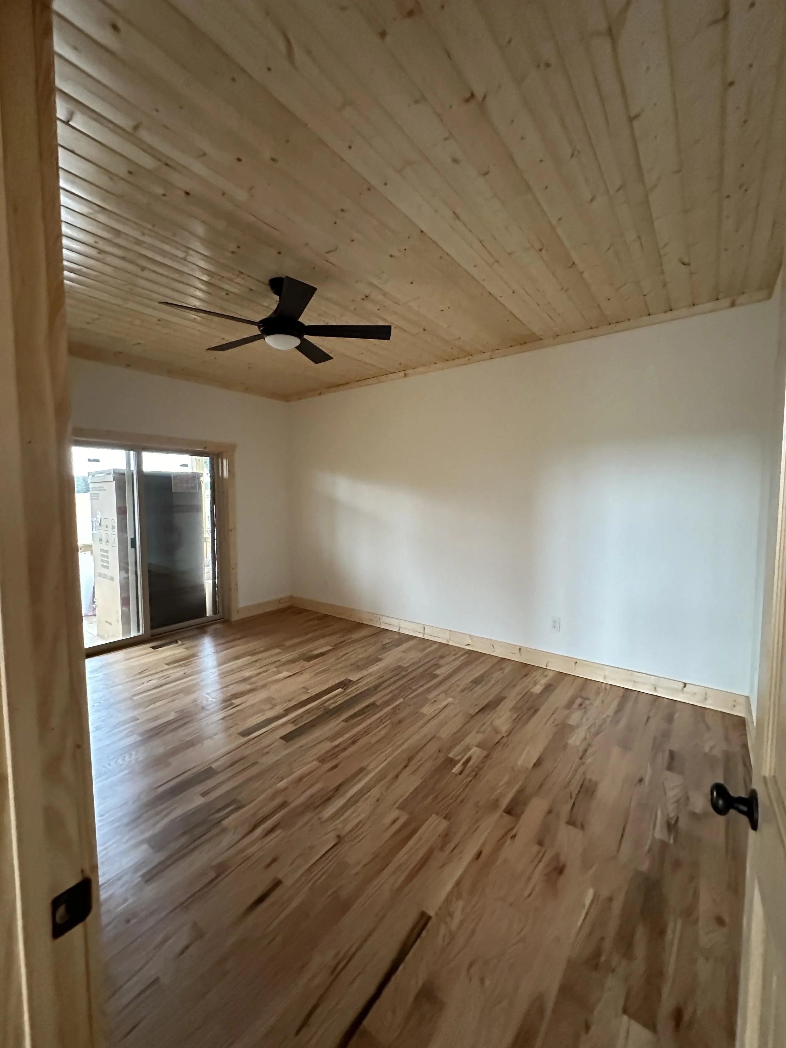 Interior view of a room with a wooden ceiling, wooden flooring, a ceiling fan, and a sliding glass door leading to an outdoor area.